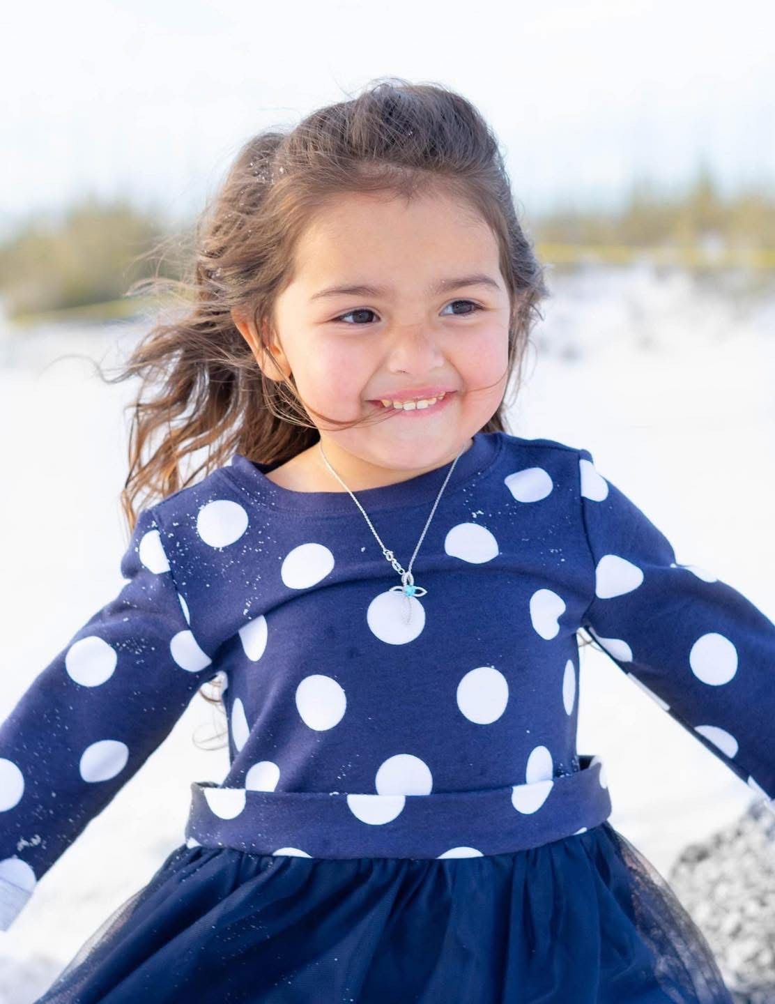 A child in a navy polka dot dress with tulle skirt plays in the snow on a bright winter day.