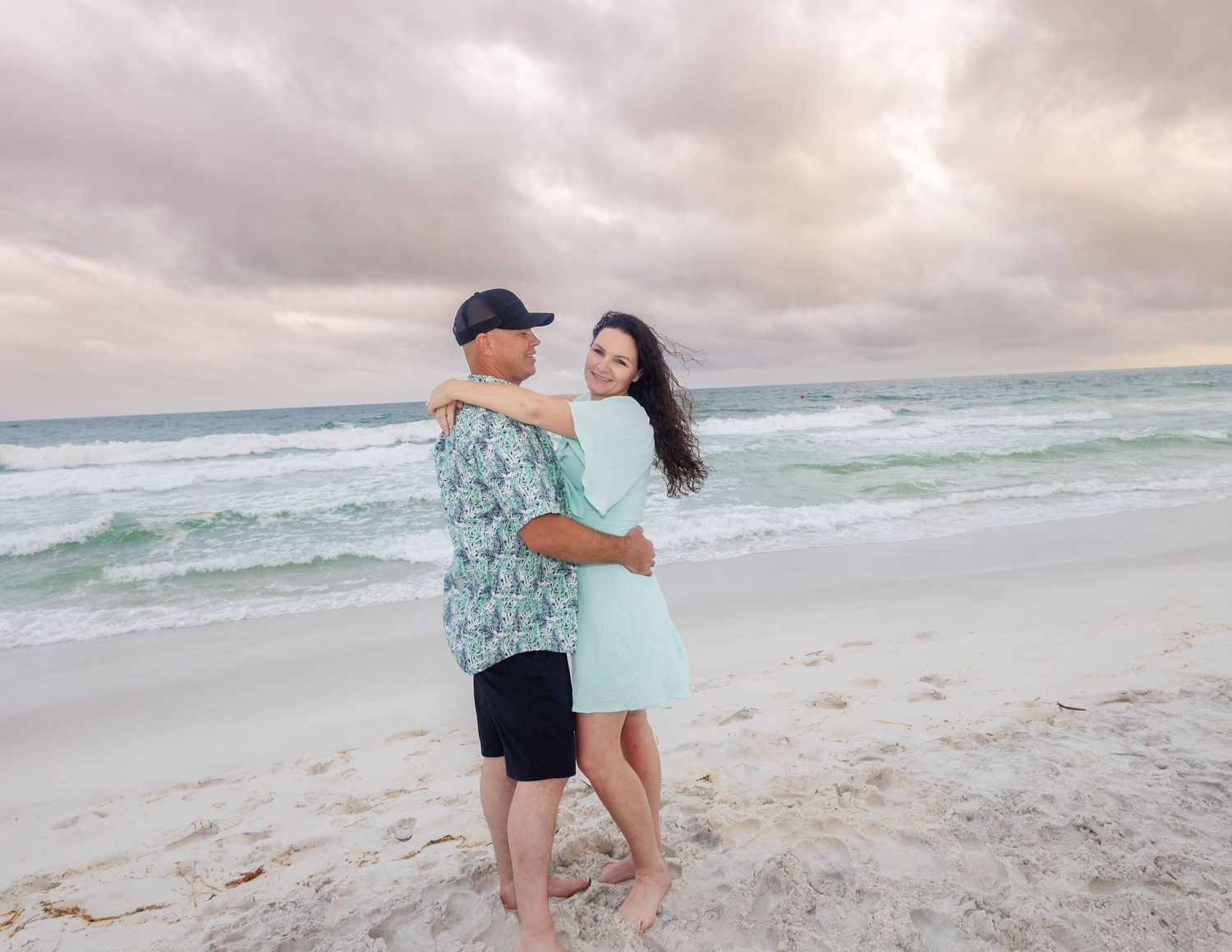 Couple embracing on a sandy beach at sunset with cloudy skies and ocean waves in the background.