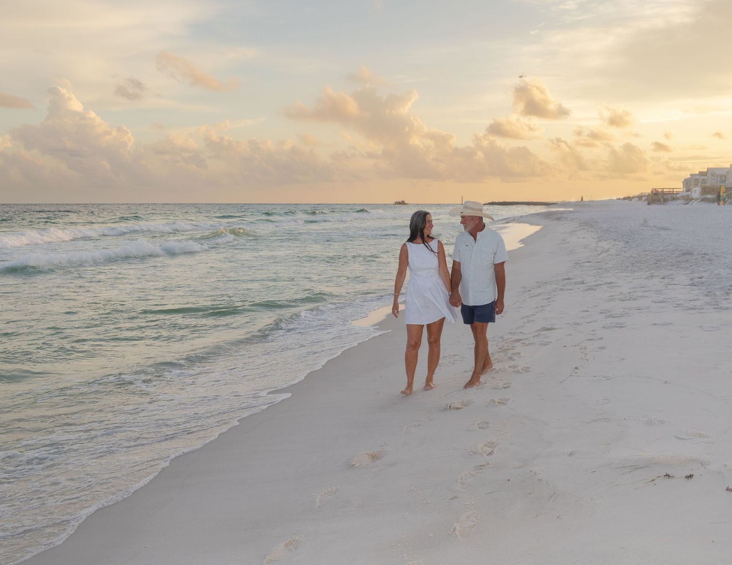 A couple in white outfits walks along a pristine beach at sunset with gentle waves rolling onto the shore.