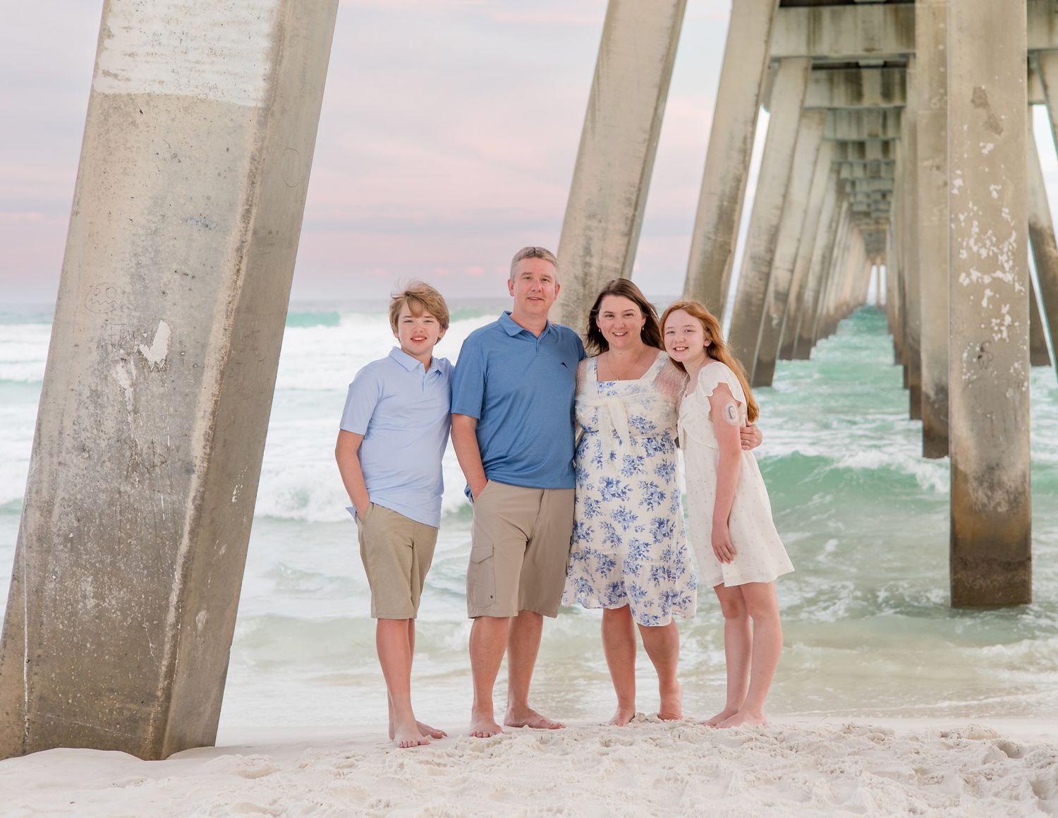 A family of four poses under a pier at the beach during sunset with ocean waves visible.