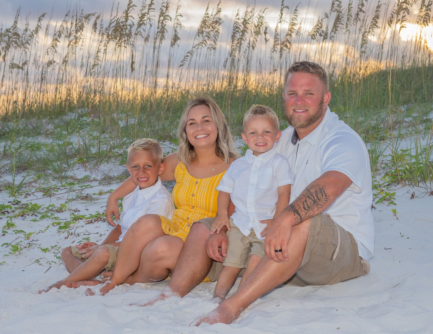 A family sits together on white sand dunes with sea oats swaying in the golden sunset light at the beach.