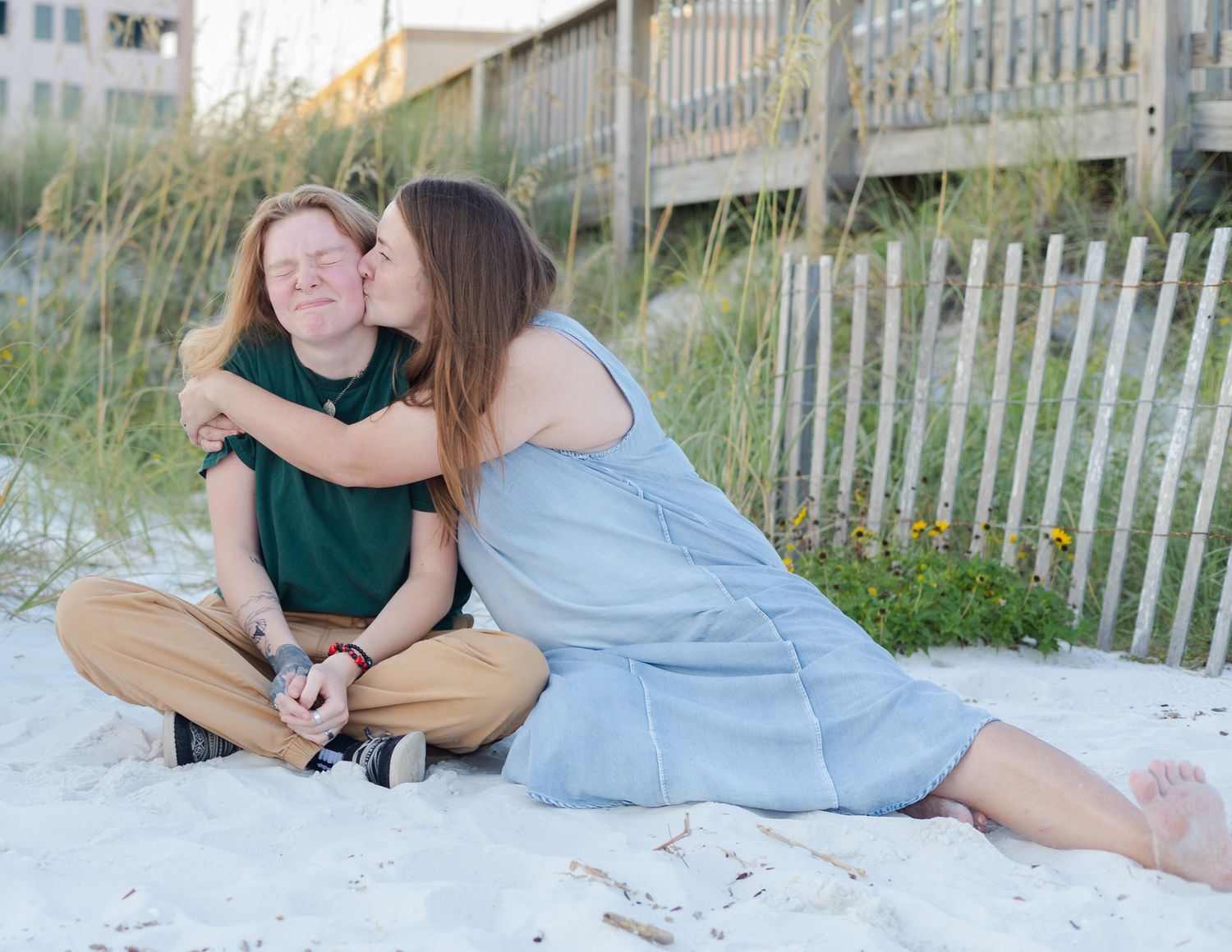 A couple in coordinated green and blue outfits poses together on a sandy beach with wooden fence in background.
