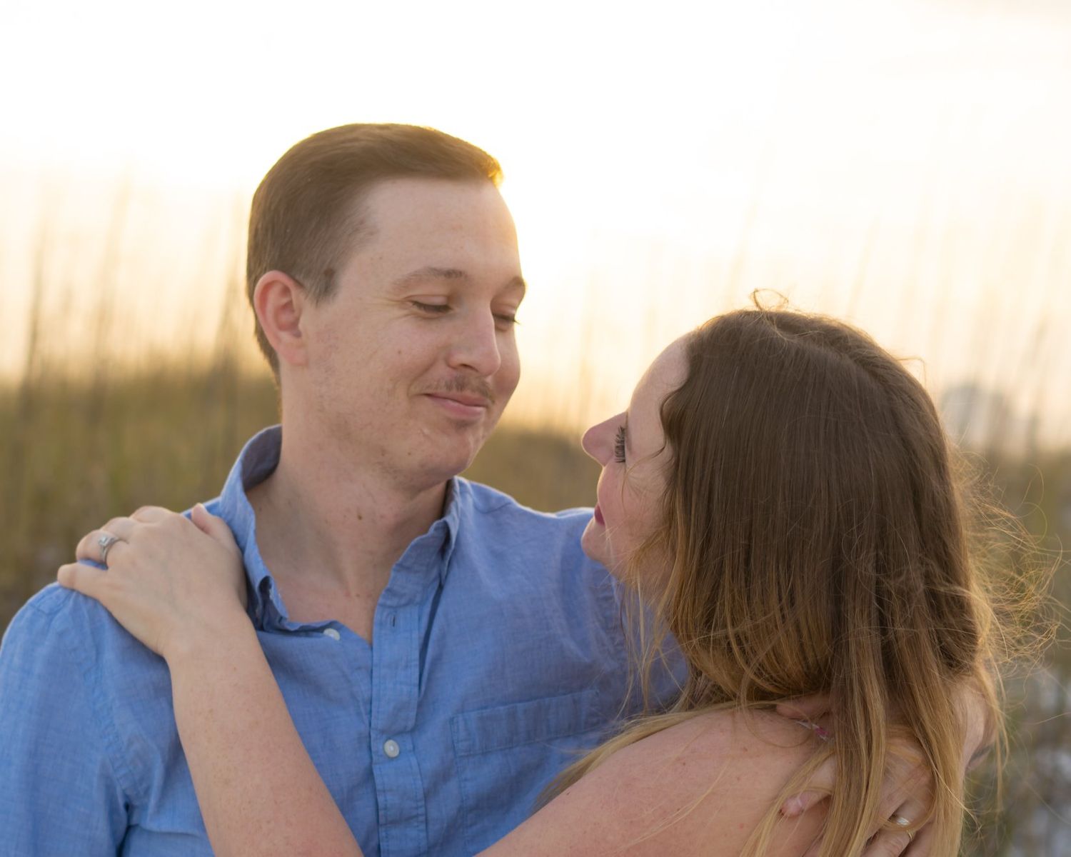 A couple in blue and white outfits share tender moments during a sunset photoshoot in a field.
