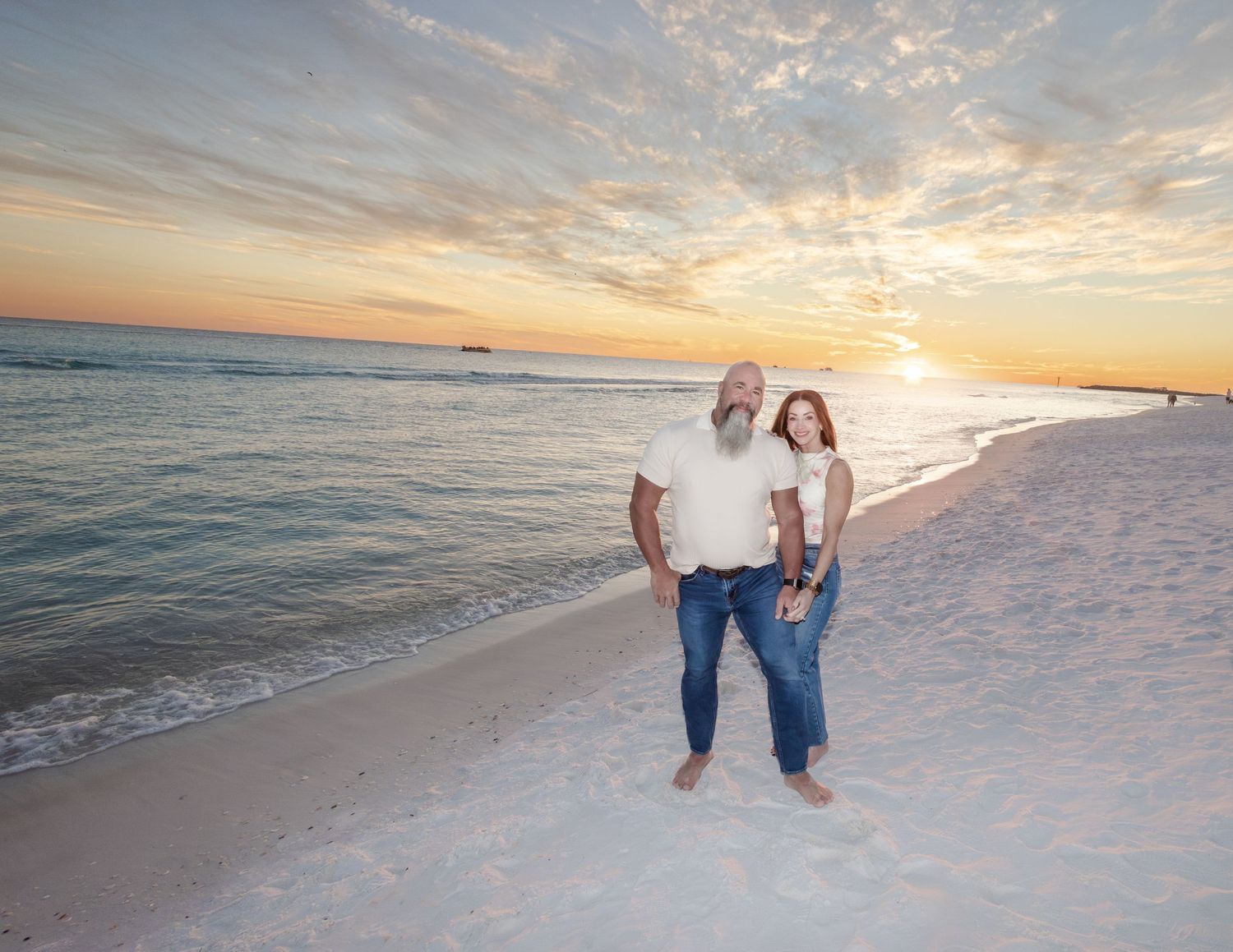 A couple stands together on a white sandy beach at sunset with gentle waves and a glowing orange sky in the background.