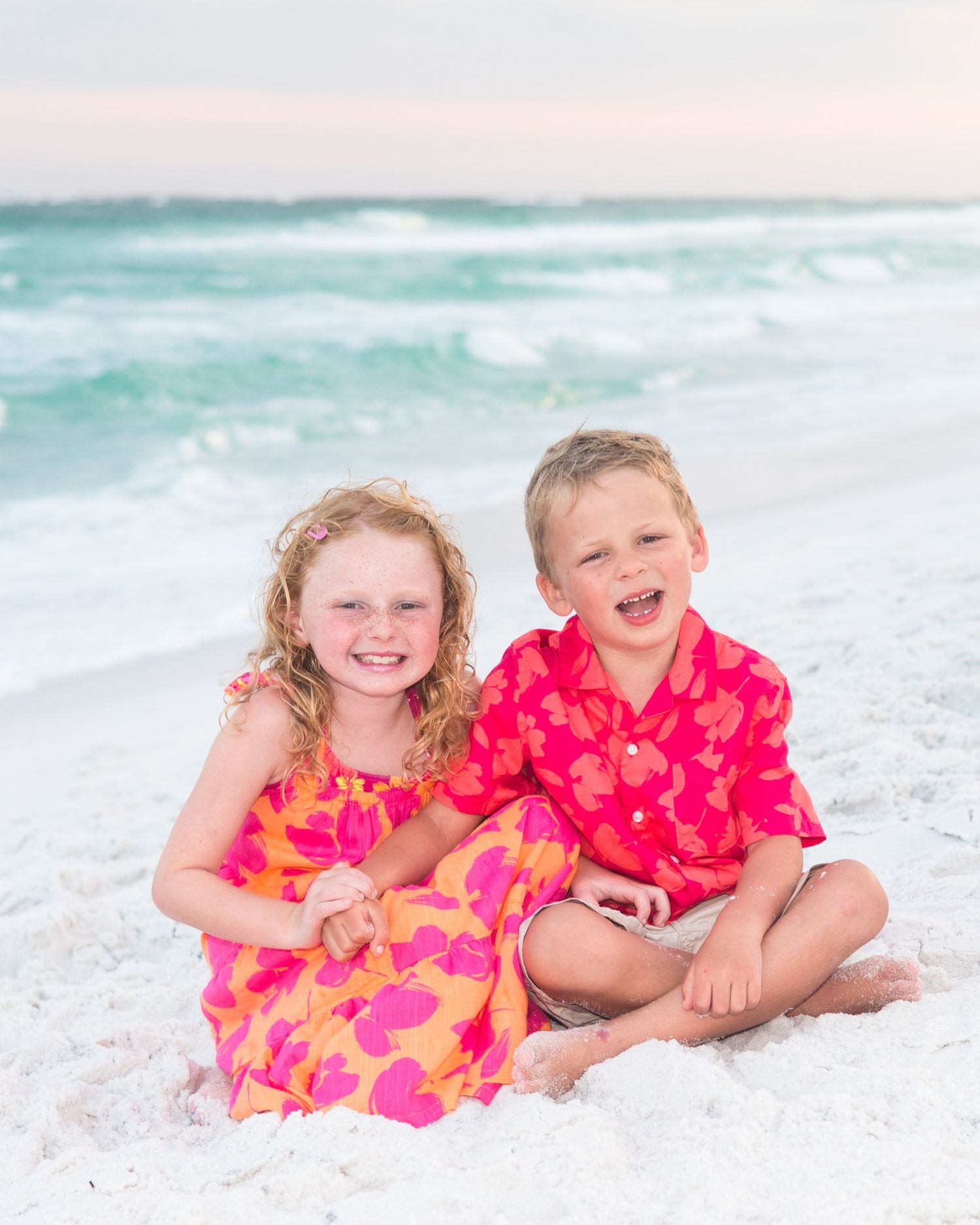 Two children wearing bright pink floral outfits play and laugh together on a white sandy beach.