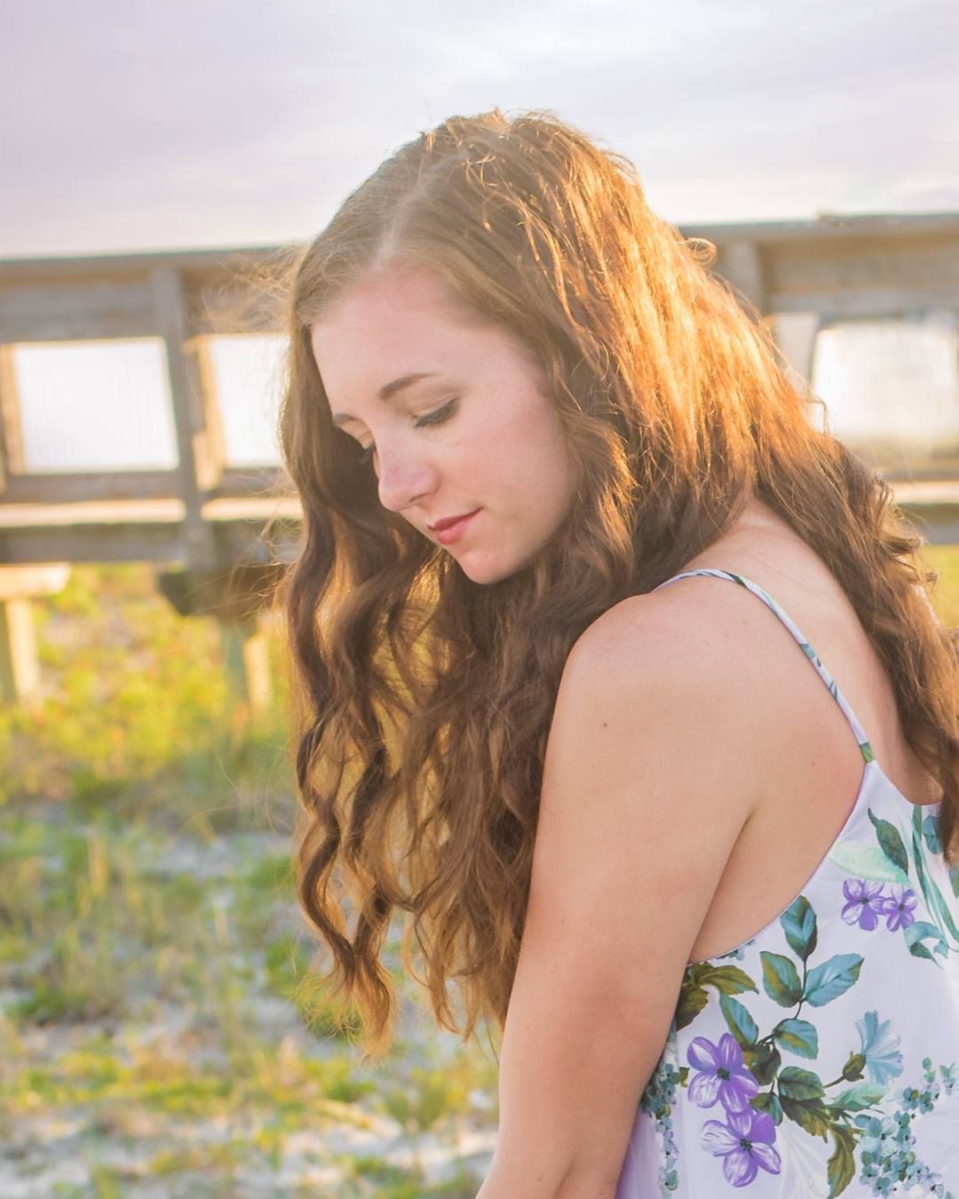 A woman in a floral dress sits in a field at sunset with long wavy hair flowing in the breeze.