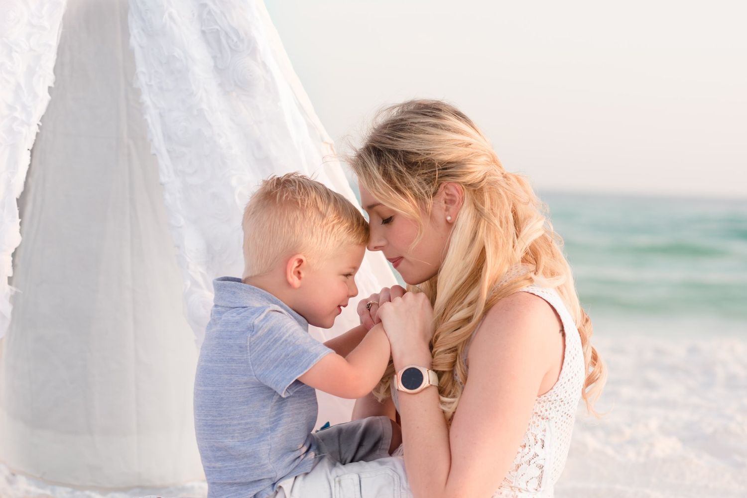 A couple shares tender moments during a beach photoshoot with soft ocean waves and white sand in the background.