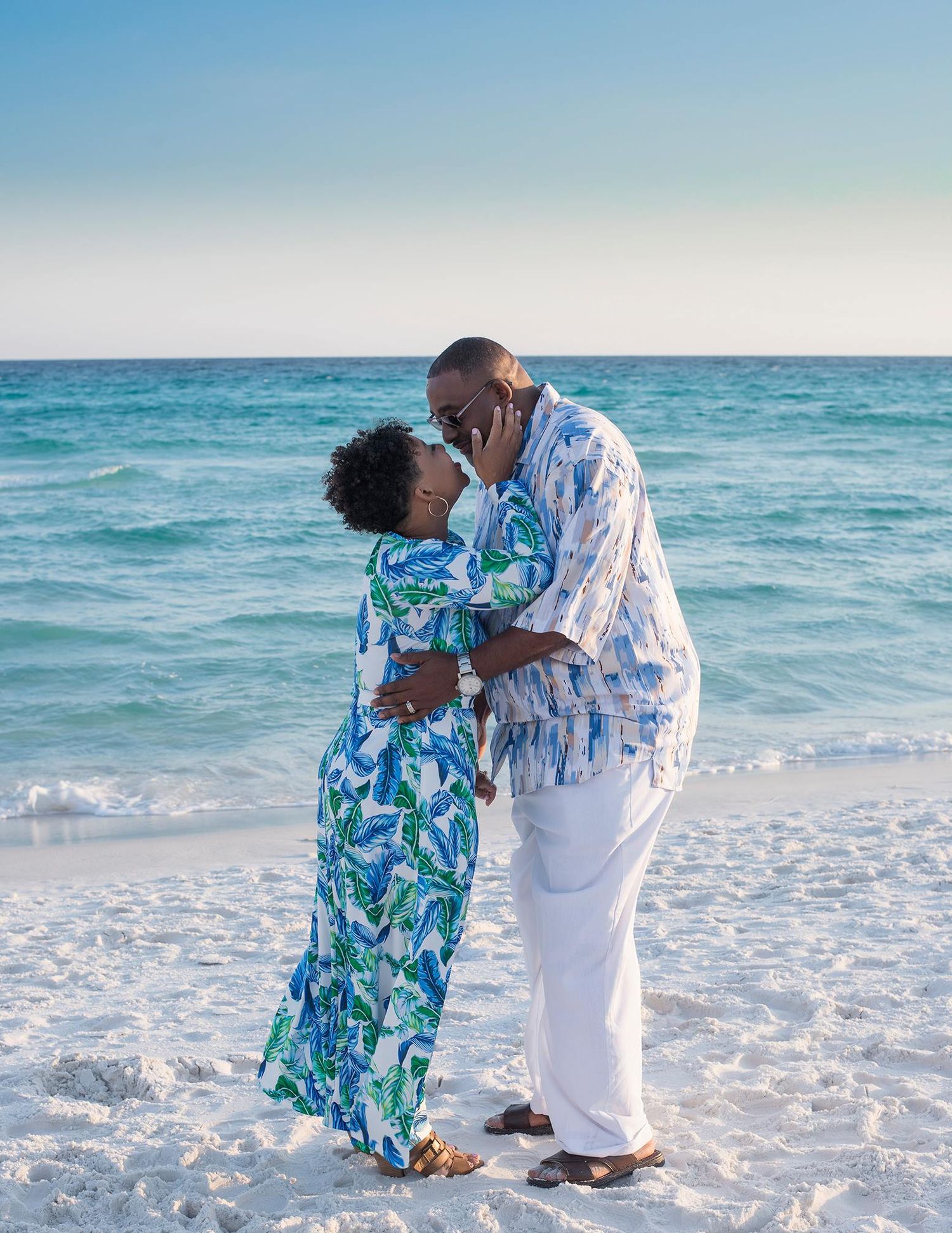 Couple in coordinating blue and white outfits sharing tender moments on a sunny beach.