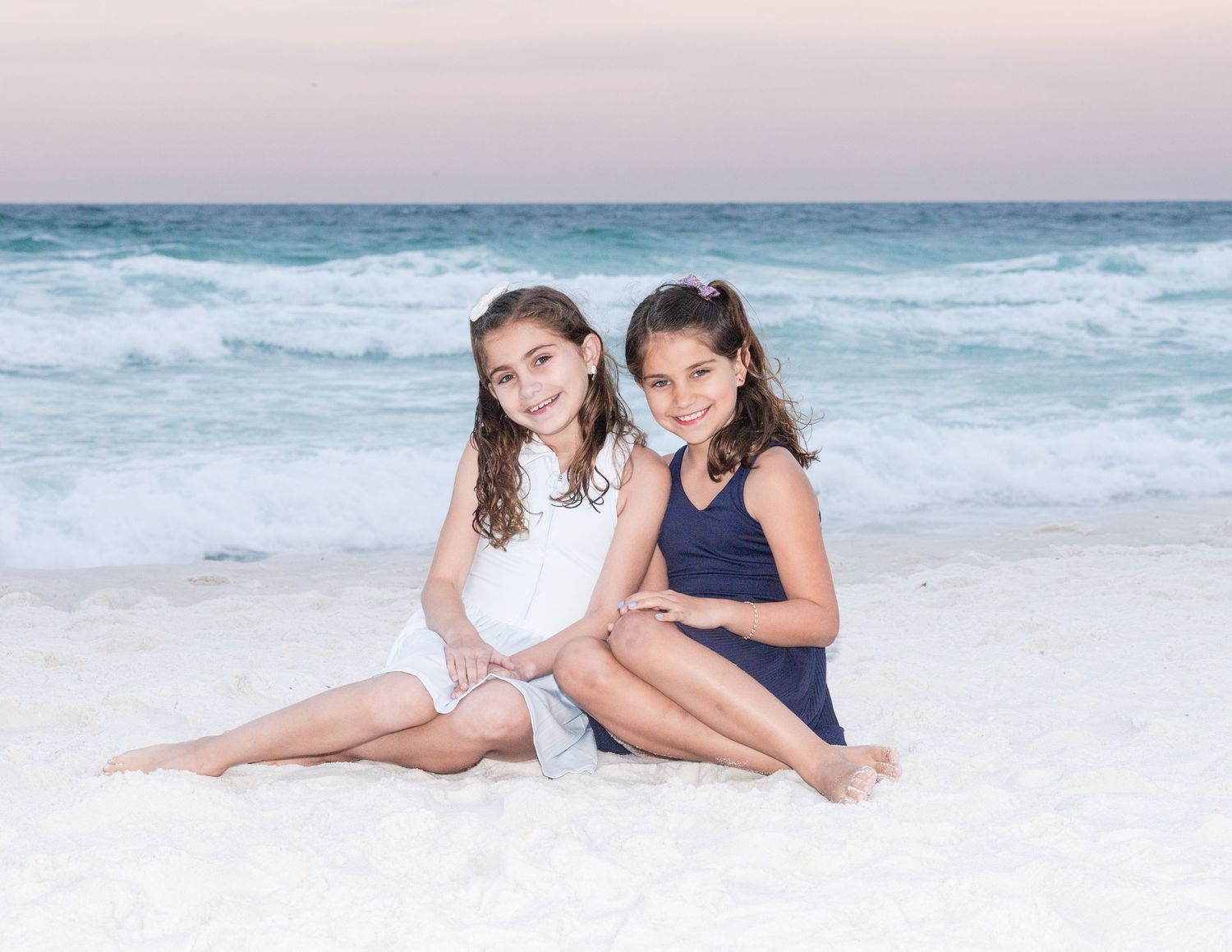Two children in navy and white outfits pose together on a sandy beach with ocean waves in the background.