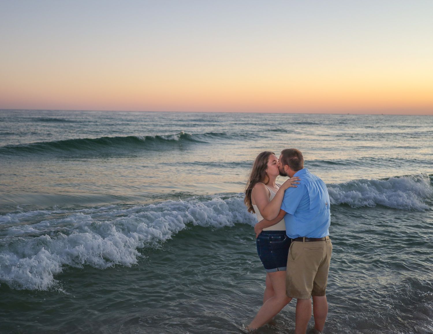 Couple embraces on a sandy beach at sunset with rolling waves in the background.