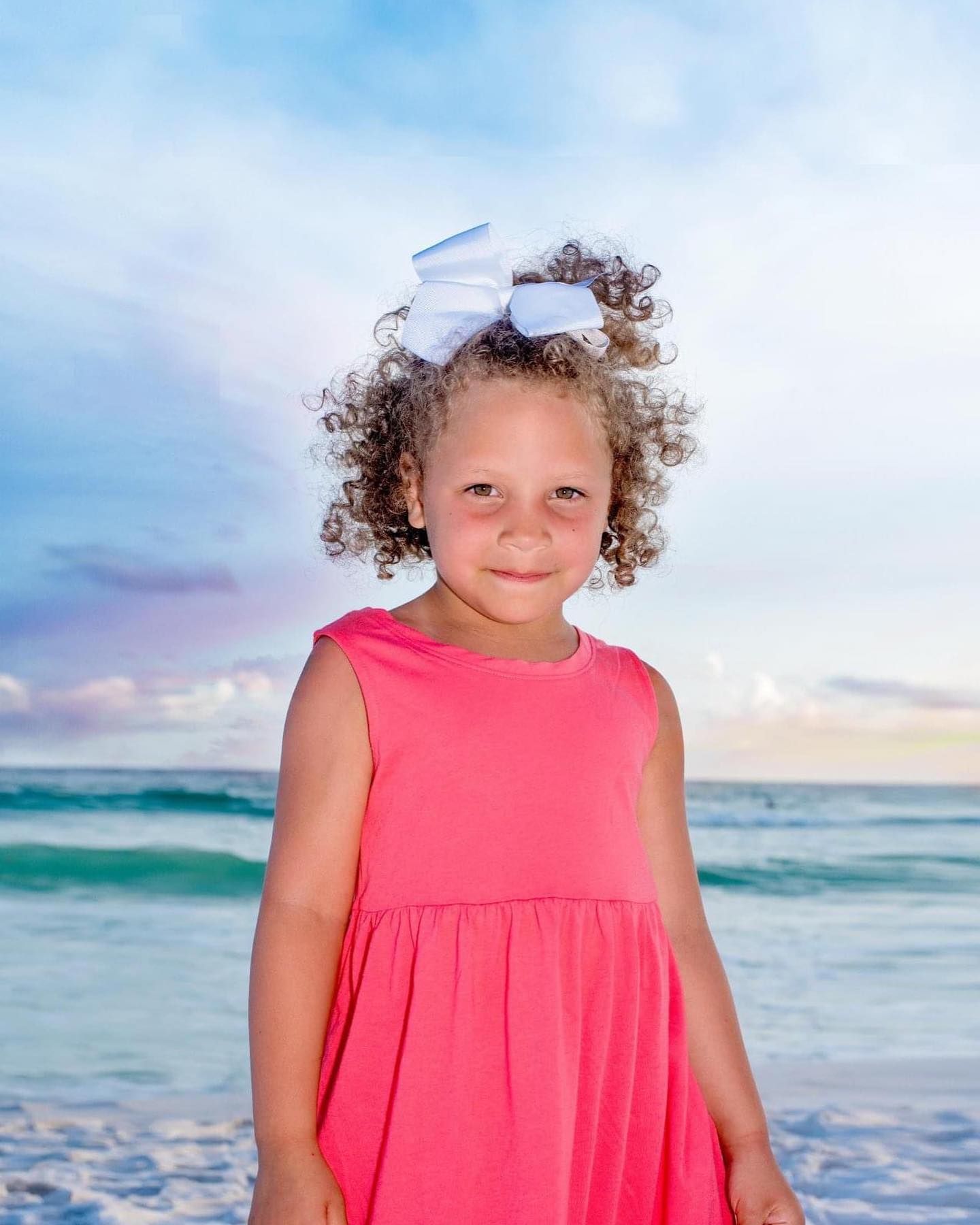 Young child in a coral pink dress plays on a beautiful beach with ocean waves in the background.