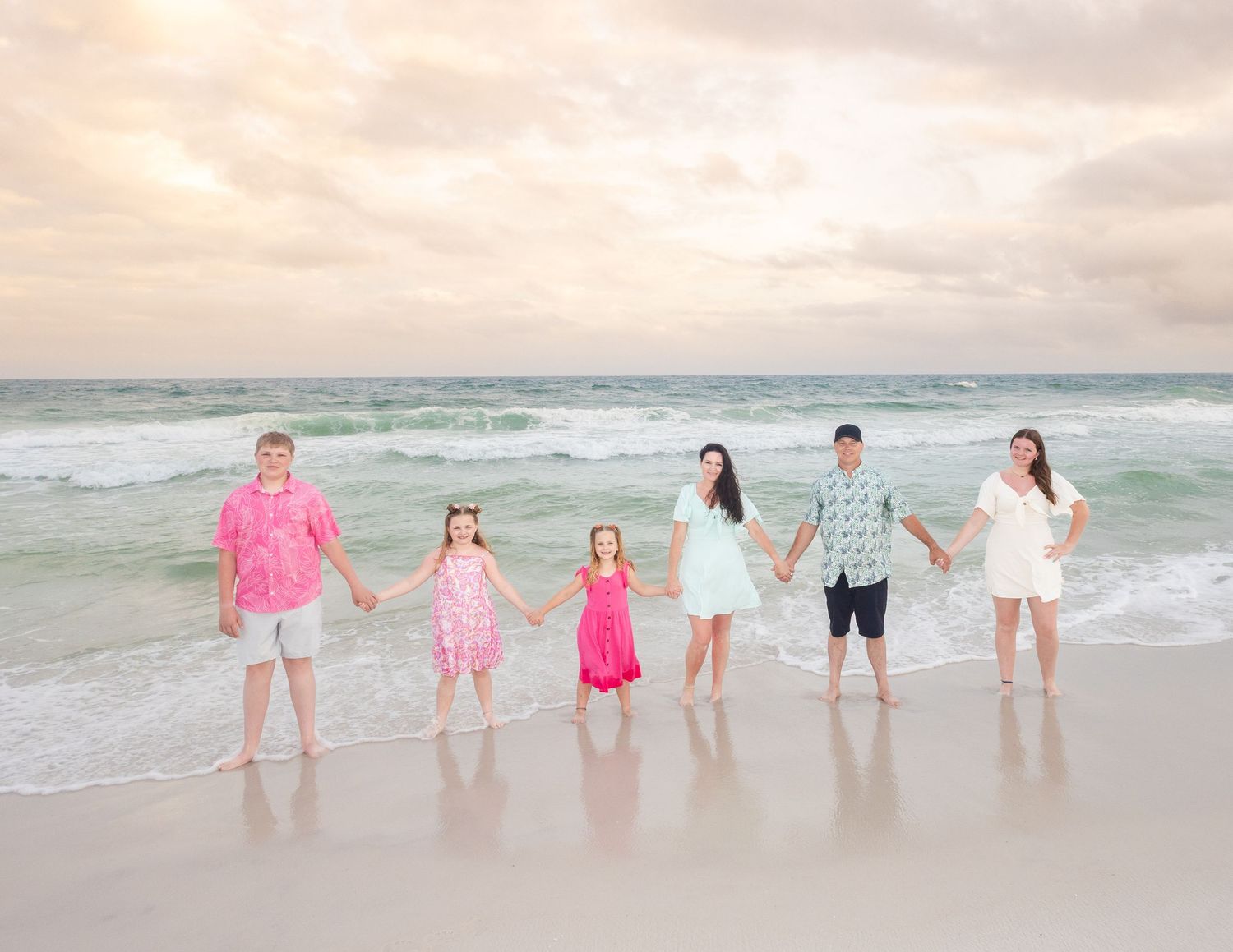 Family of six holding hands on a sandy beach at sunset, wearing colorful summer outfits with ocean waves behind them.