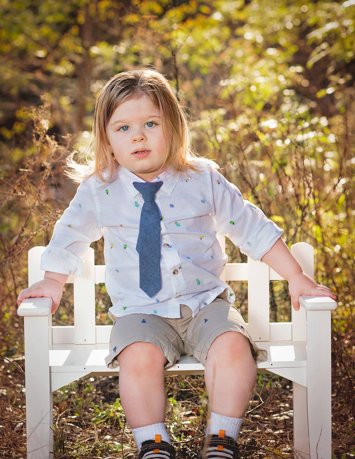 A child in a white shirt and blue tie sits on a white wooden bench in a golden autumn setting.