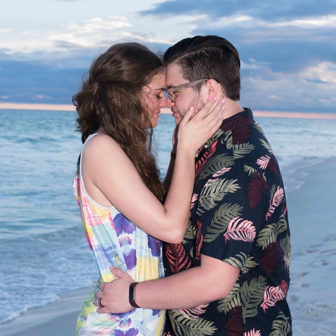 A couple embraces and kisses on a beach at sunset with the ocean waves in the background.