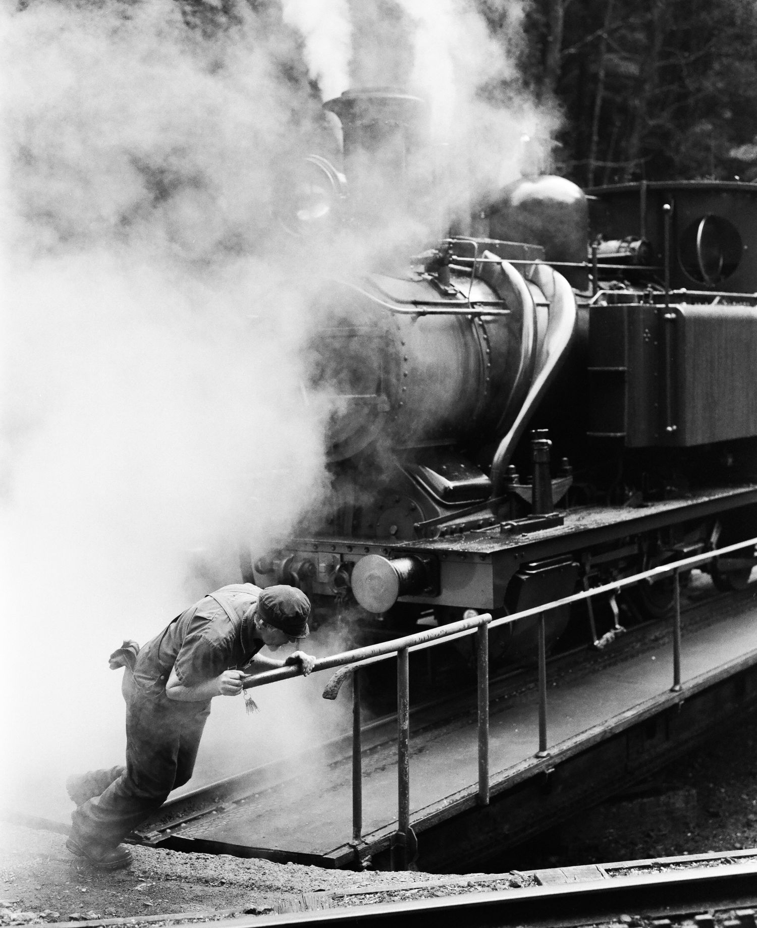 Steam billows from vintage locomotive while worker performs maintenance on railway.