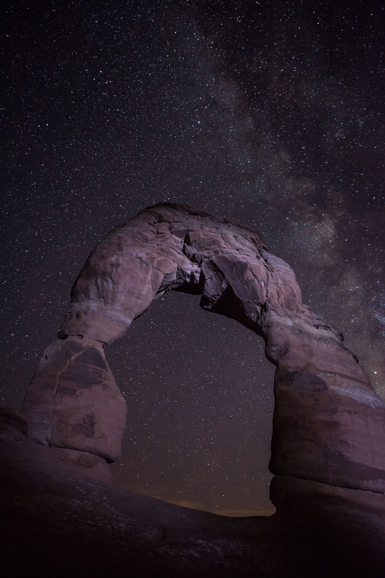 Natural stone arch formation in Arches National Park illuminated under a starry night sky with the Milky Way visible.