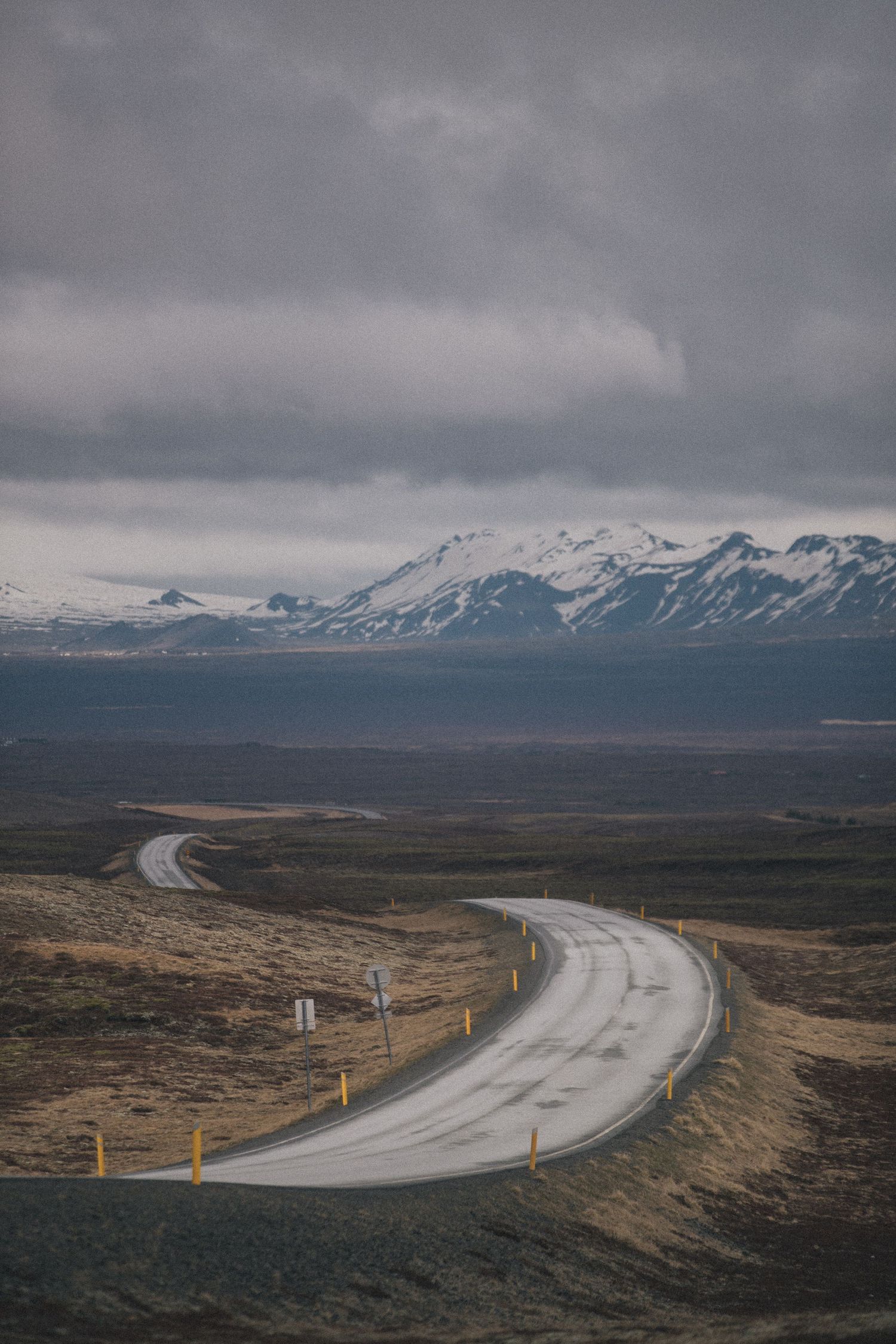 Winding mountain road leads through barren landscape toward snow-capped peaks under stormy skies.
