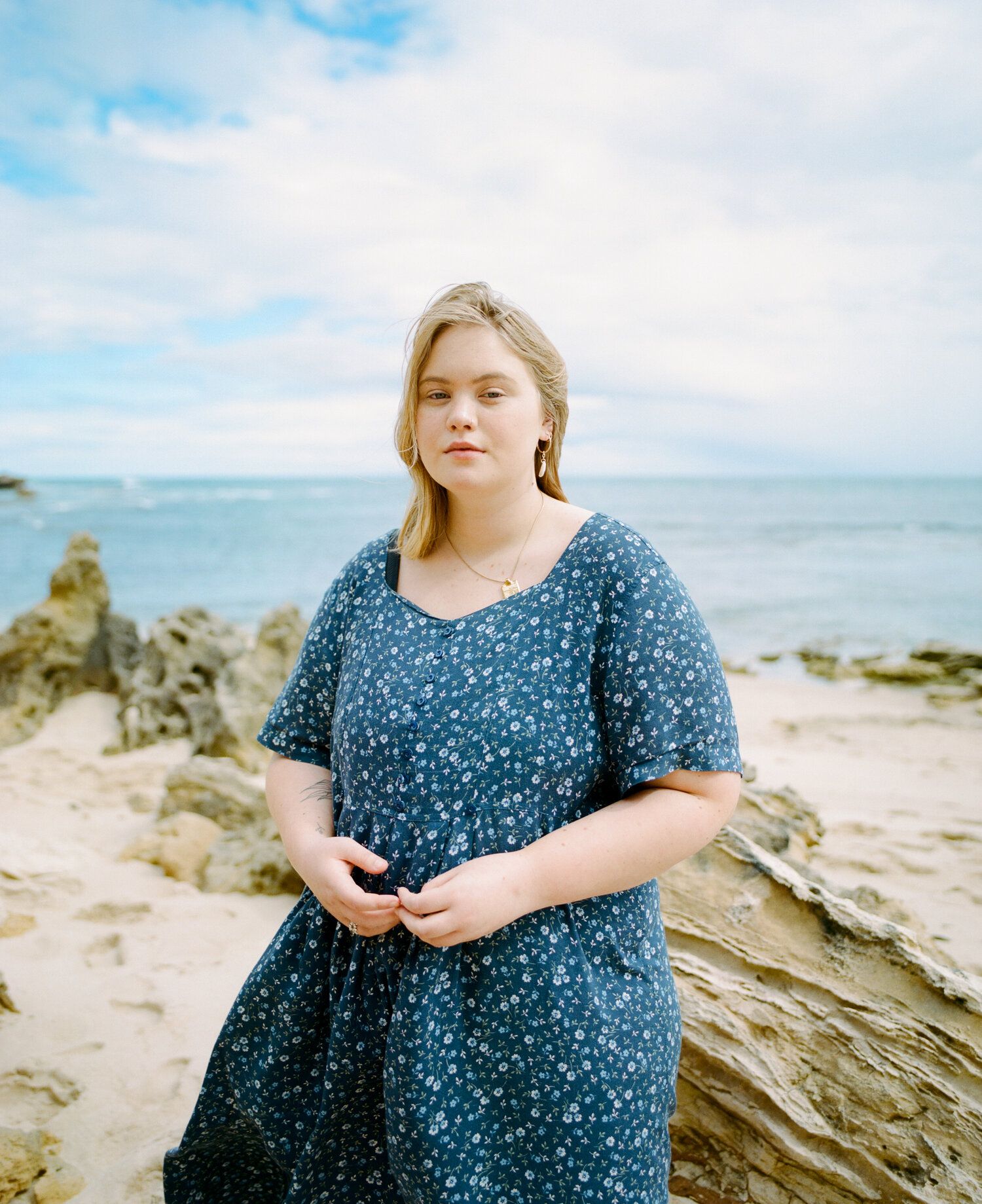 A person in a blue polka dot dress stands on a rocky beach with the ocean and cloudy sky in the background.