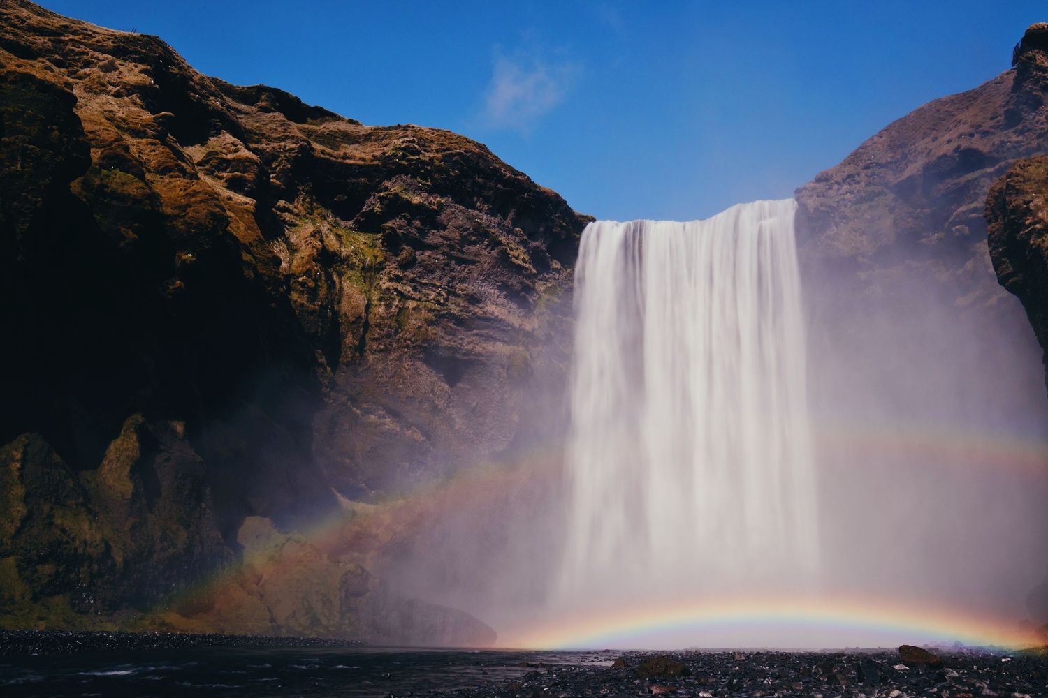 Majestic waterfall cascades down rocky cliffs with a vivid rainbow appearing in the mist against a blue sky.