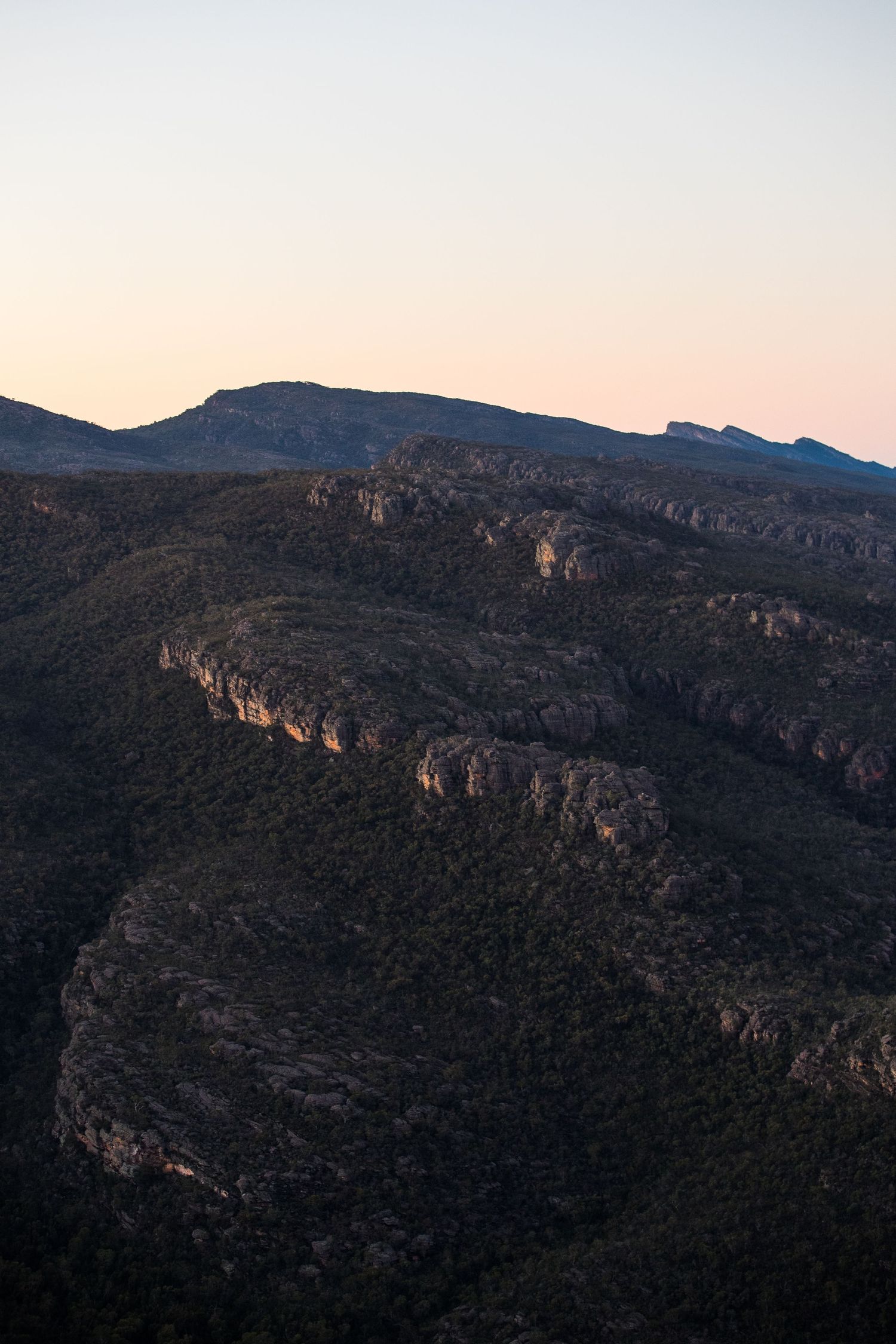 Rugged mountain ridges cast dramatic shadows at dusk with layered rocky cliffs against a pale pink sky.
