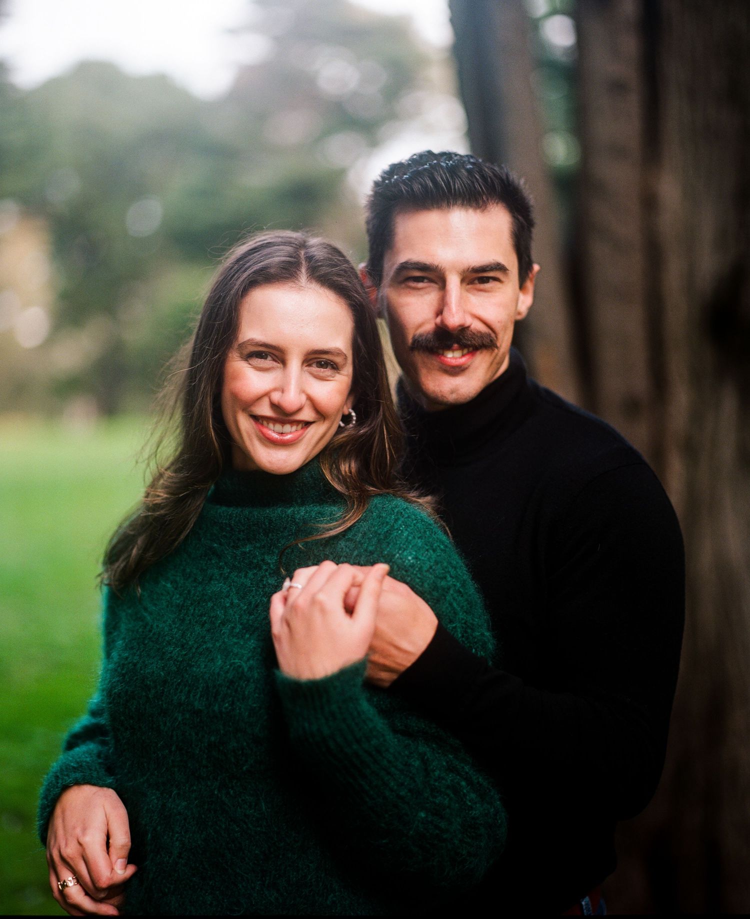 Couple embracing outdoors with woman in emerald sweater and man in black turtleneck in natural setting.