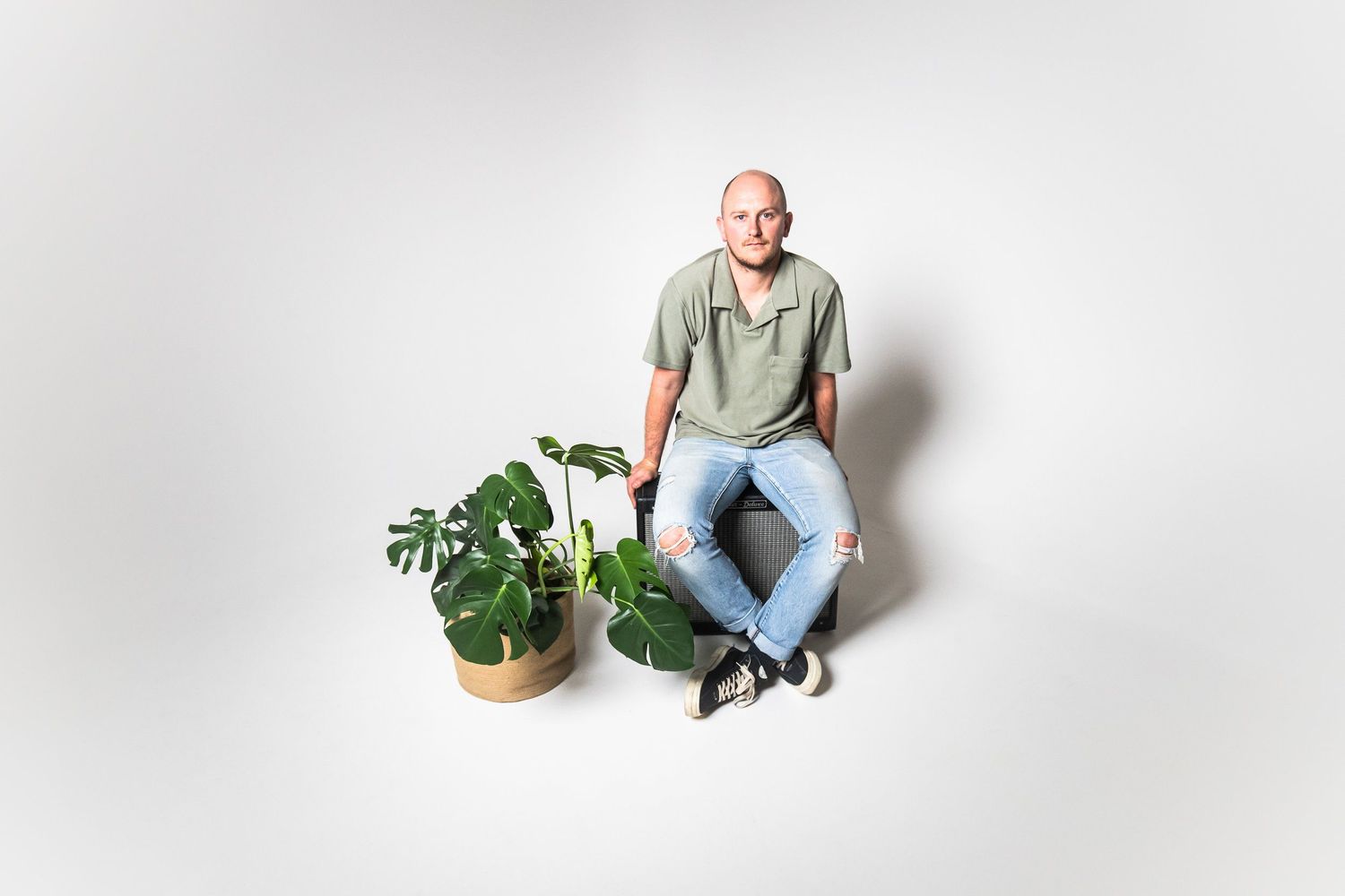 Person sits casually between potted plants against white background in minimalist portrait.