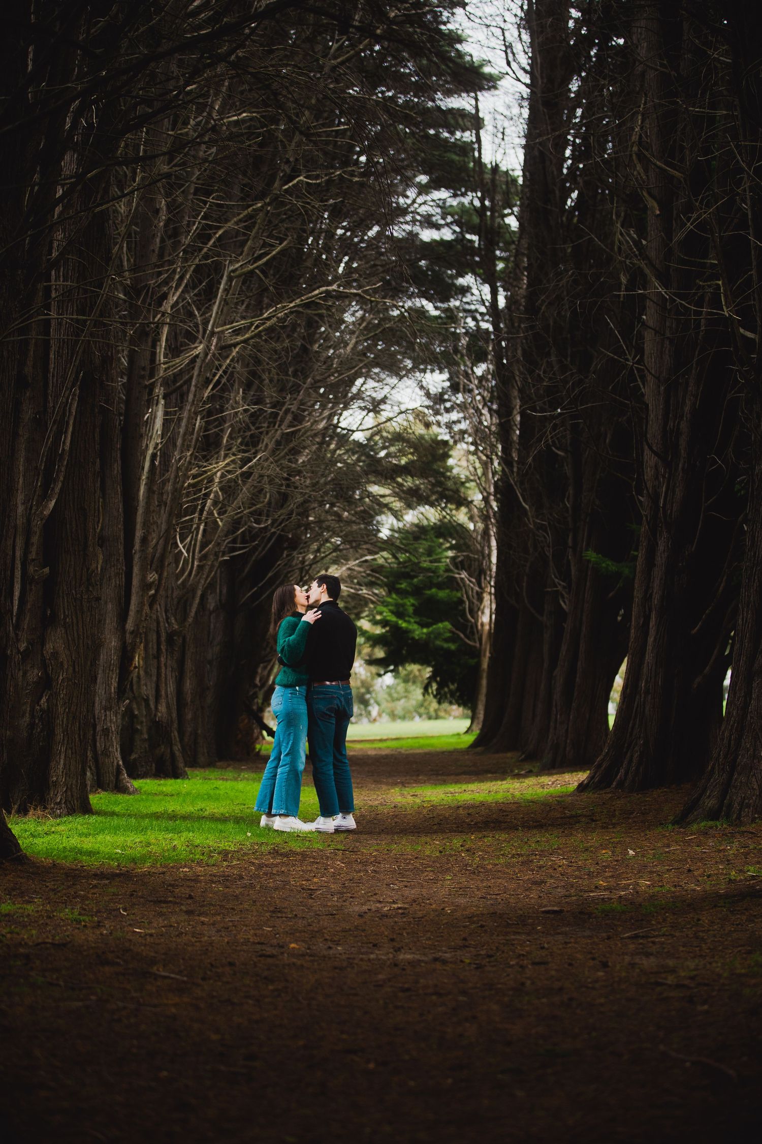 Two people embrace while standing on a dirt path beneath towering cypress trees forming a natural tunnel.