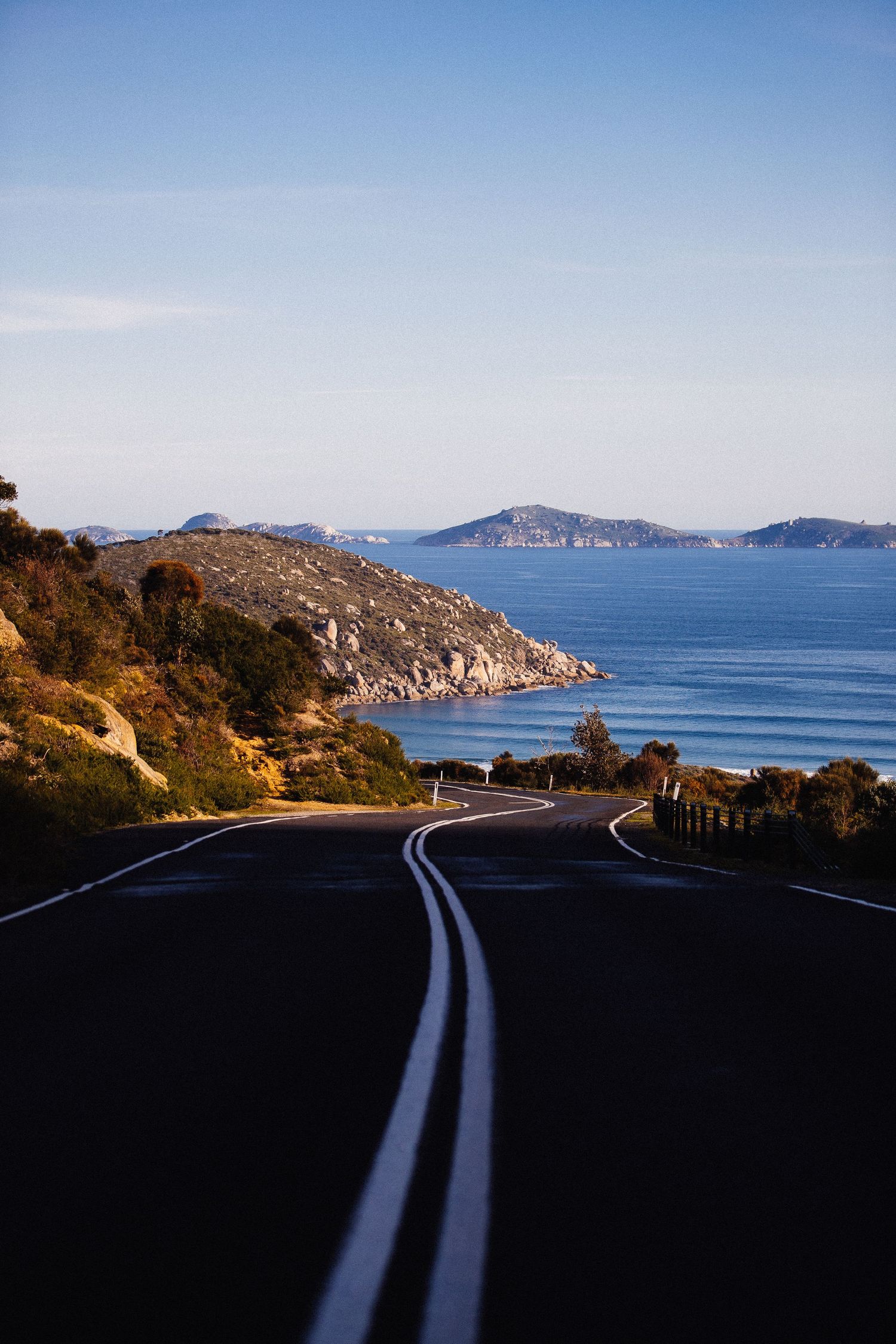Winding coastal road curves along rocky cliffs overlooking a serene blue ocean bay.