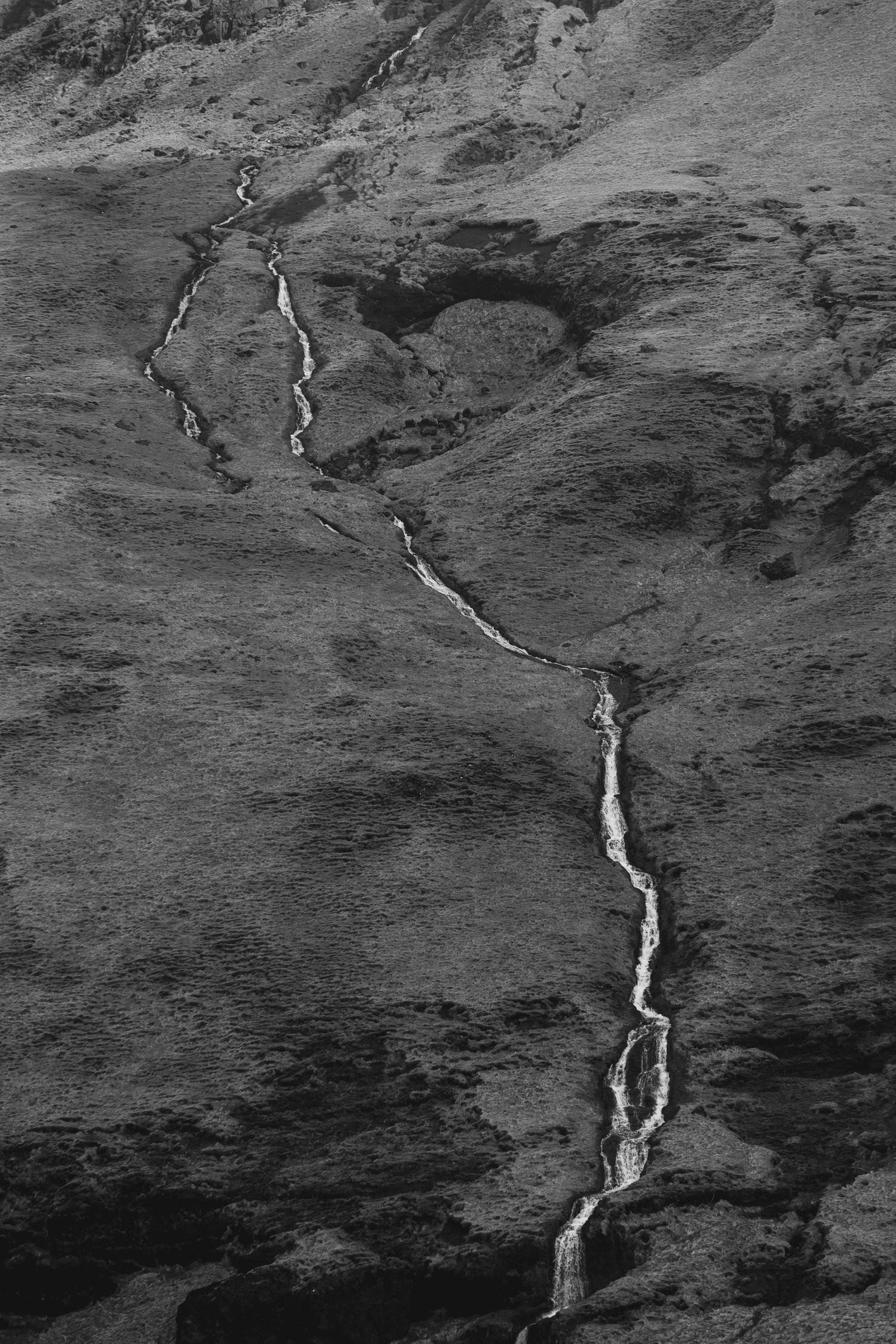 A black and white aerial view of a mountain stream winding and cascading down a steep hillside.