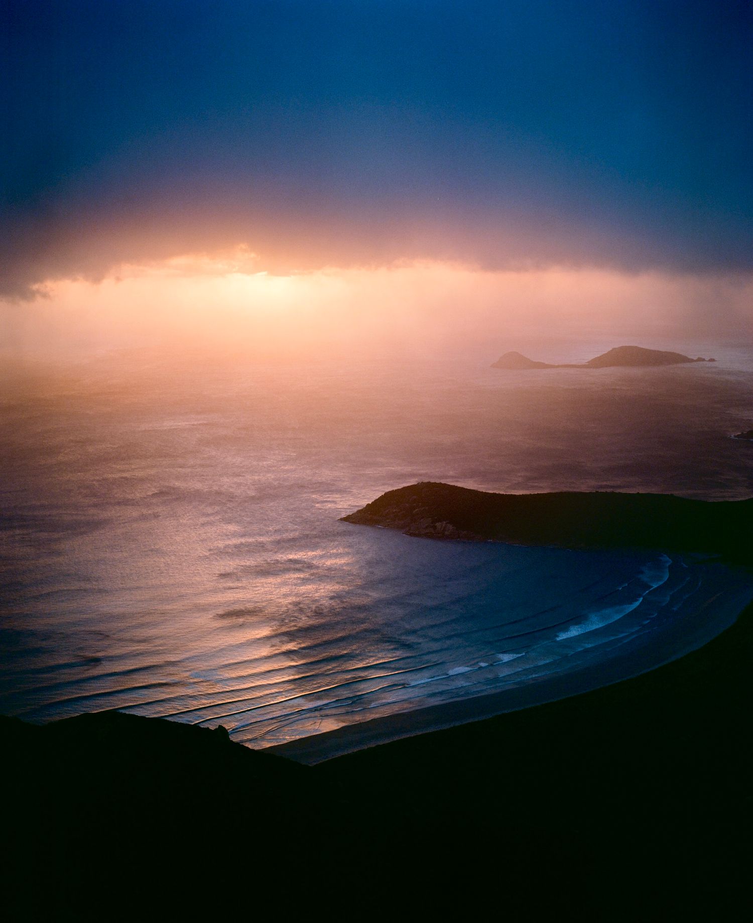 Dramatic sunset illuminates misty coastal landscape with rolling hills and curving beach along rugged shoreline.