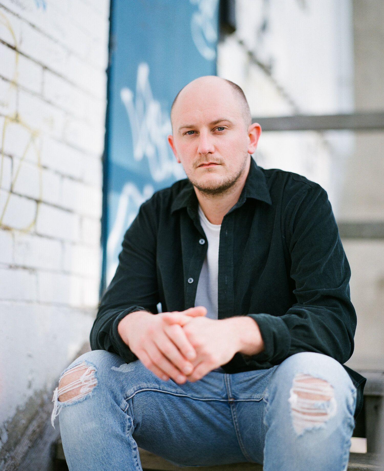 Person in black jacket and ripped jeans sitting on steps against white brick wall in casual urban portrait.