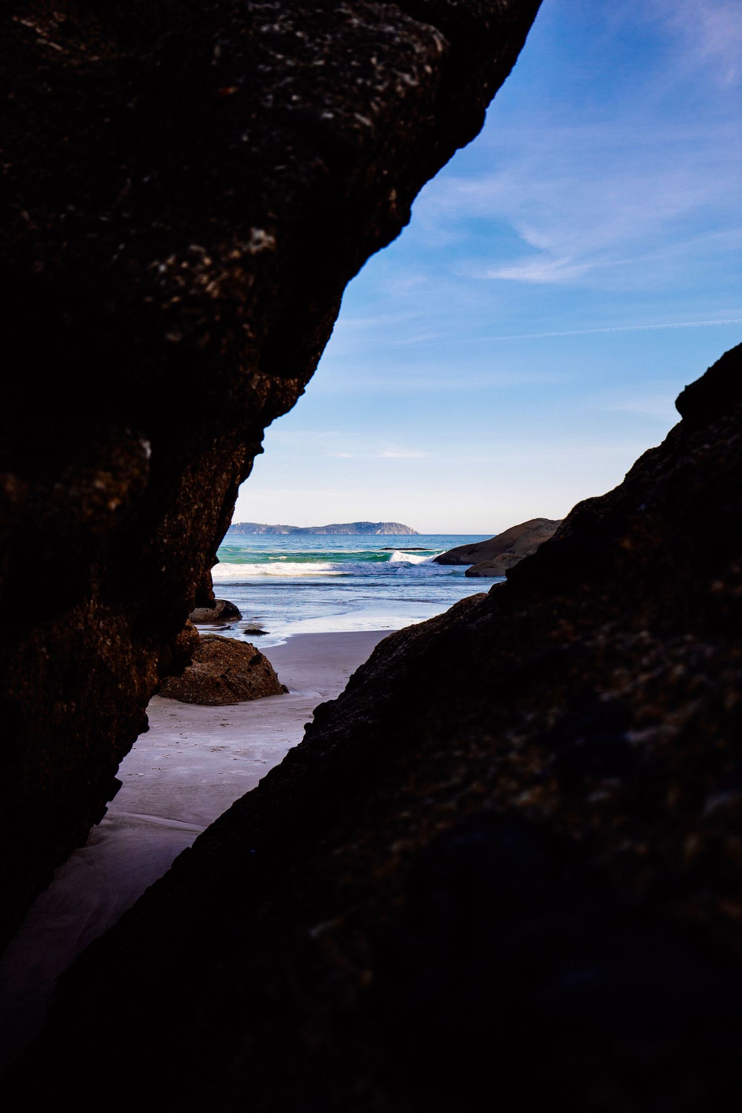 View through a rocky cave opening reveals a serene beach landscape with blue ocean and distant hills on the horizon.