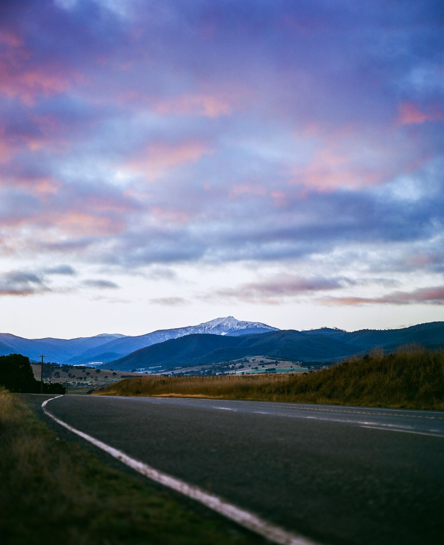 A curving mountain road at sunset with dramatic pink and purple clouds over snow-capped peaks in the distance.