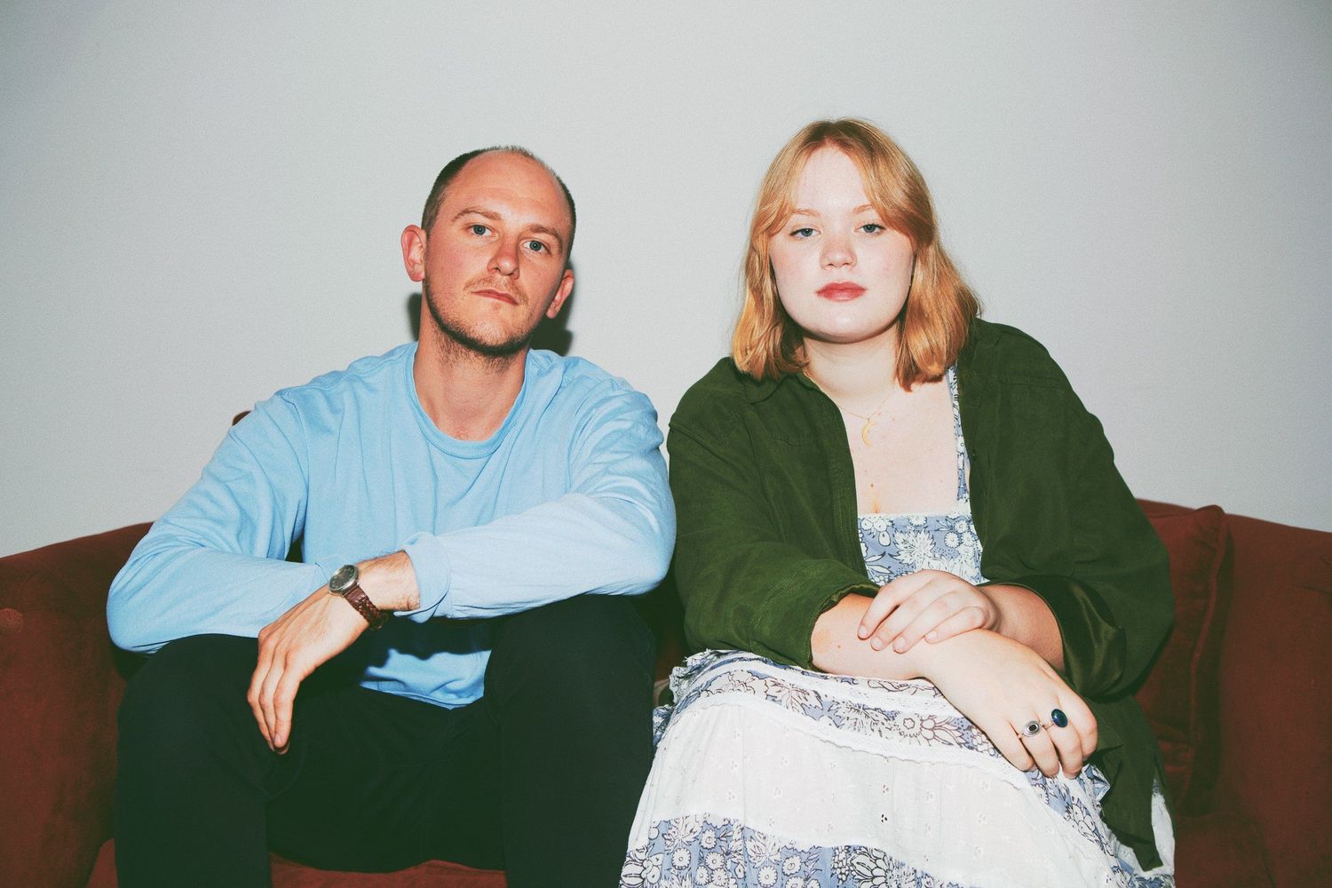 Two people sit on a dark couch, one wearing a light blue sweater and one in a green jacket.