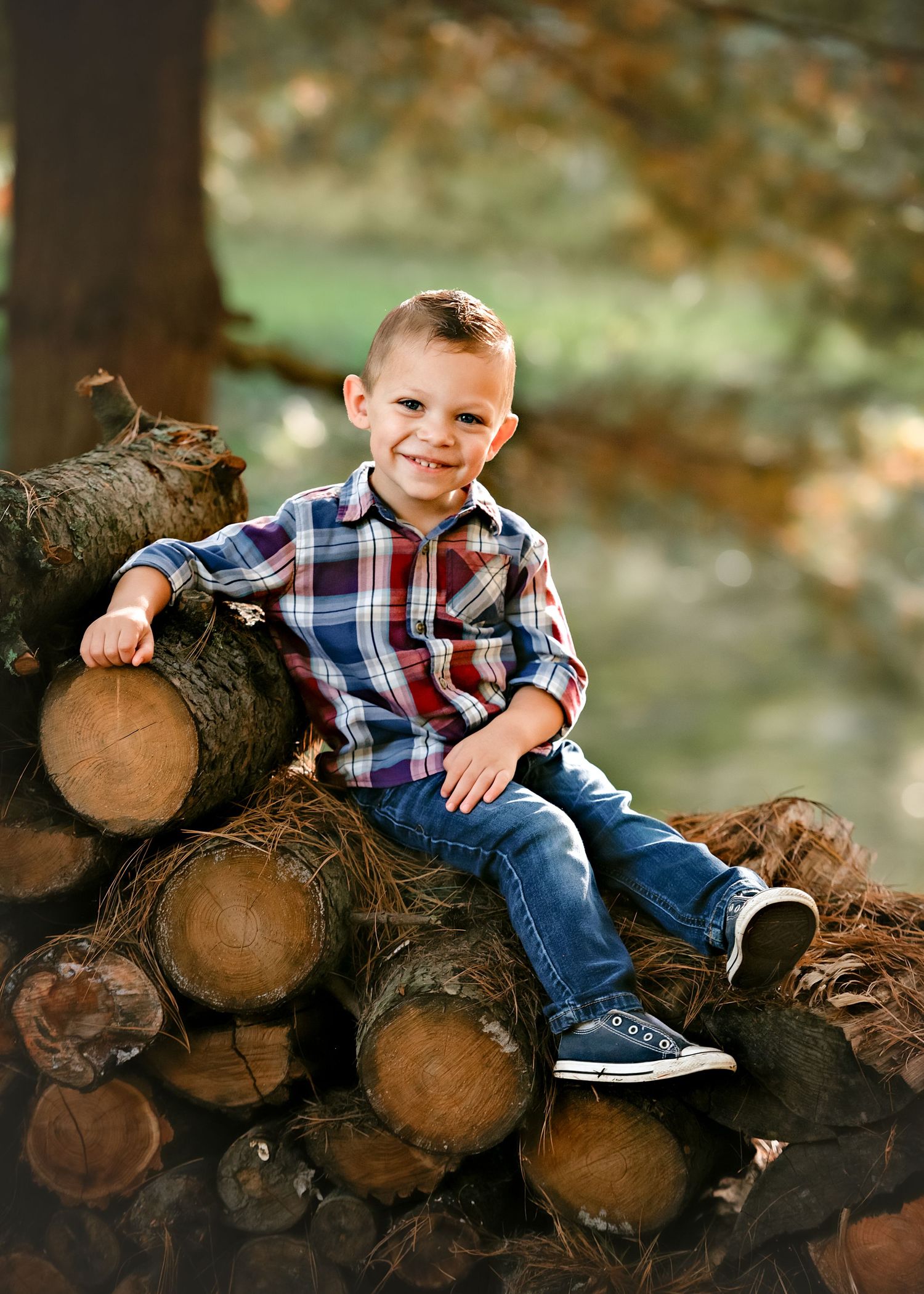 Child sitting on stacked logs in plaid shirt during autumn outdoor photoshoot.