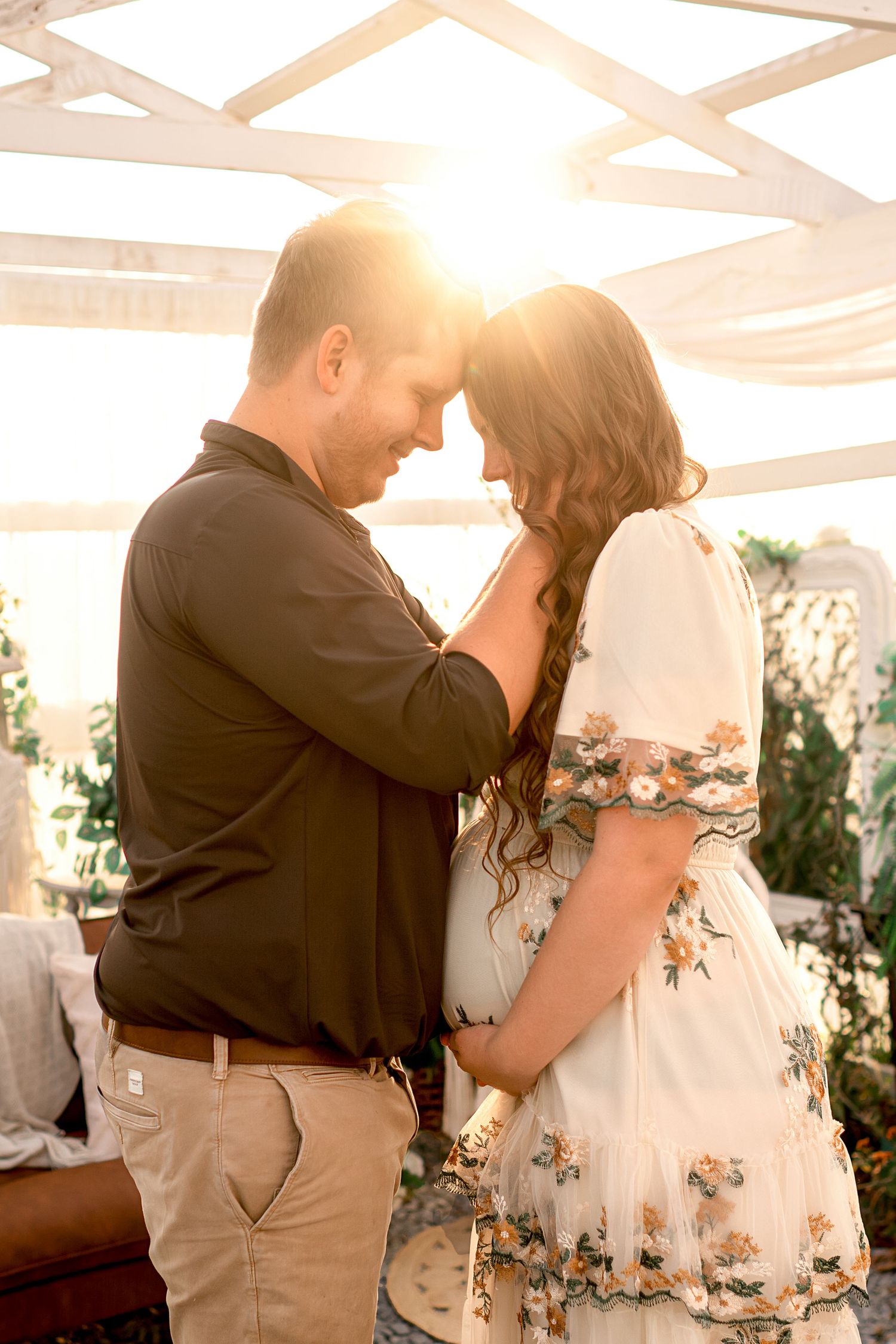 A couple shares an intimate moment under a white pergola decorated with floral arrangements at sunset.