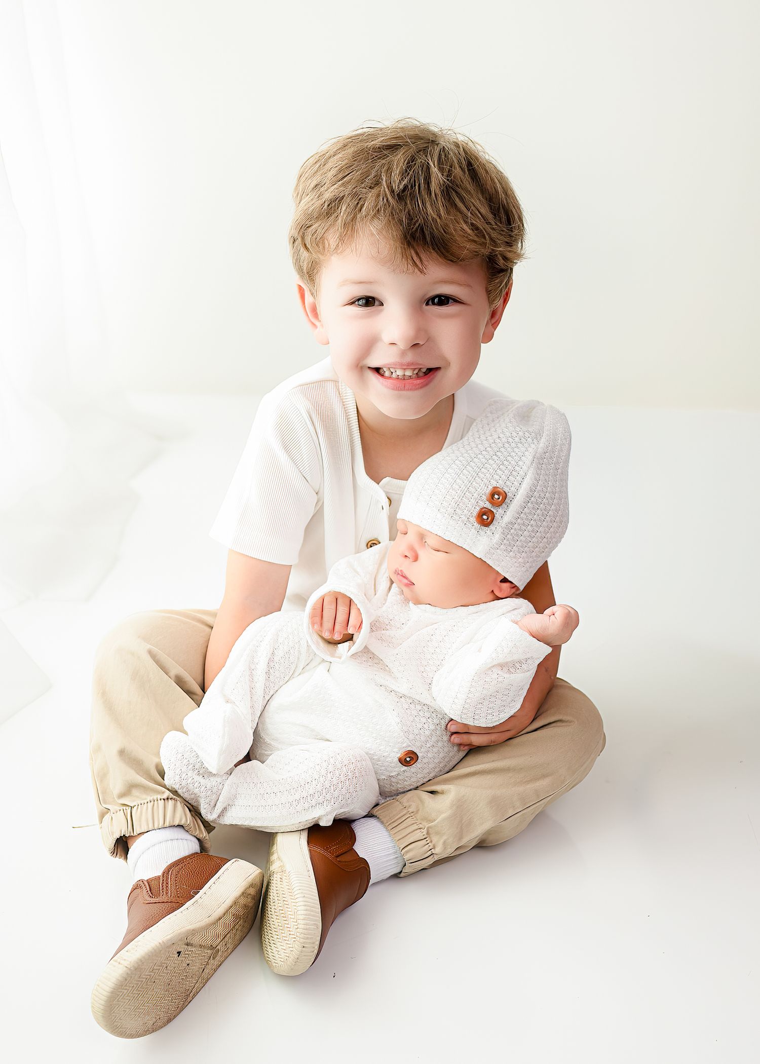 Child sitting cross-legged on white background wearing casual clothes while holding a small baby in white outfit.