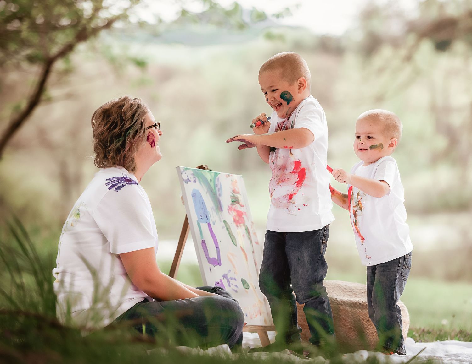 Family wearing white clothes plays with colorful powder during an outdoor photo session in a dreamy natural setting.