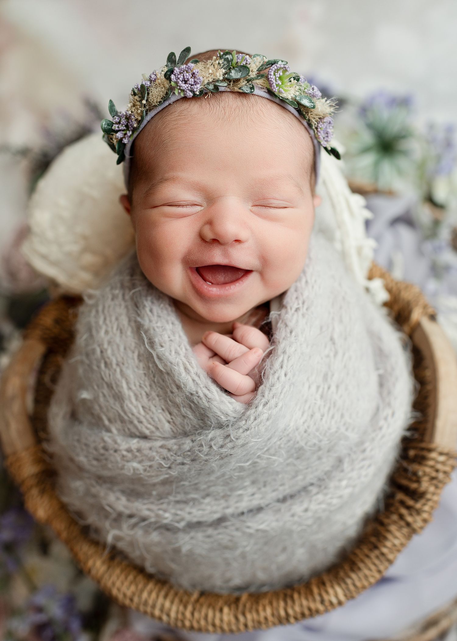 A newborn wrapped in soft white swaddle sleeps peacefully in a woven basket with delicate floral crown.
