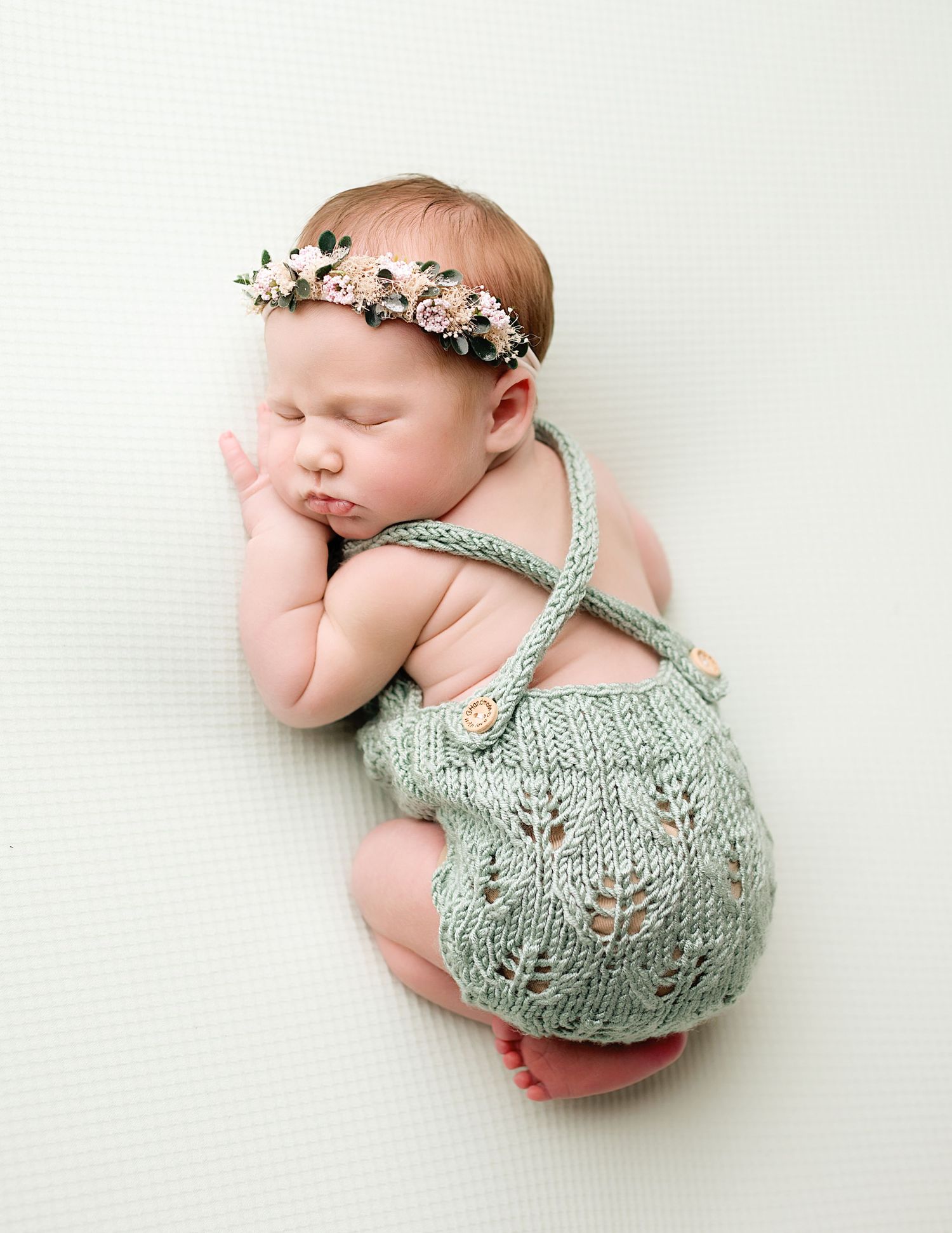 Newborn baby wearing mint green knit romper and floral crown sleeping on white backdrop.
