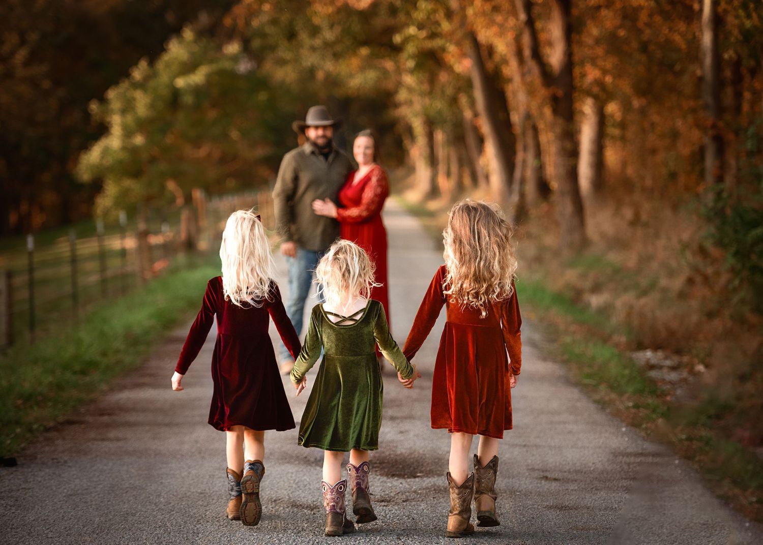 Three children in velvet dresses and boots walk down a tree-lined path in autumn.