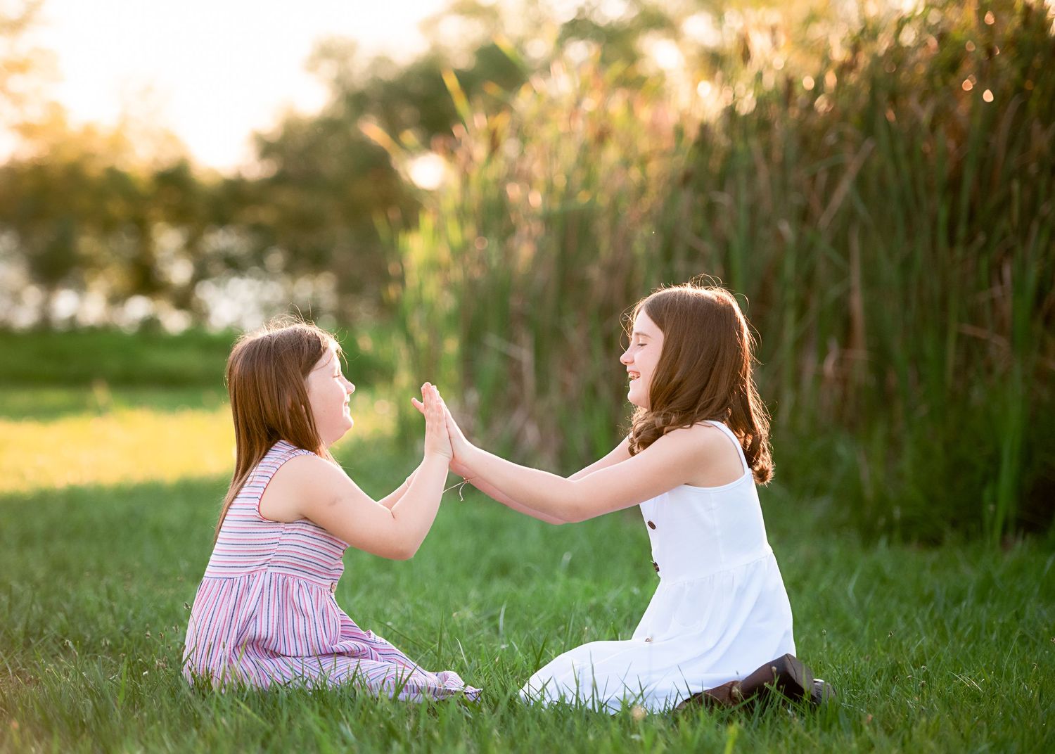 Two people sitting in a grassy field reaching out to touch hands at sunset.