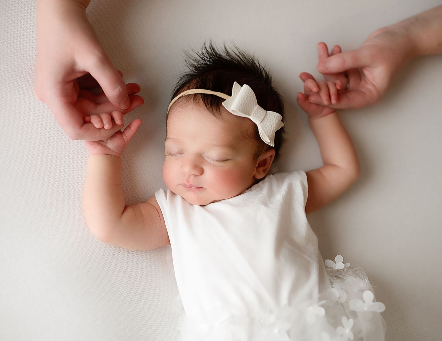 A newborn baby in a white dress sleeps peacefully on a soft white background in a gentle, artistic composition.