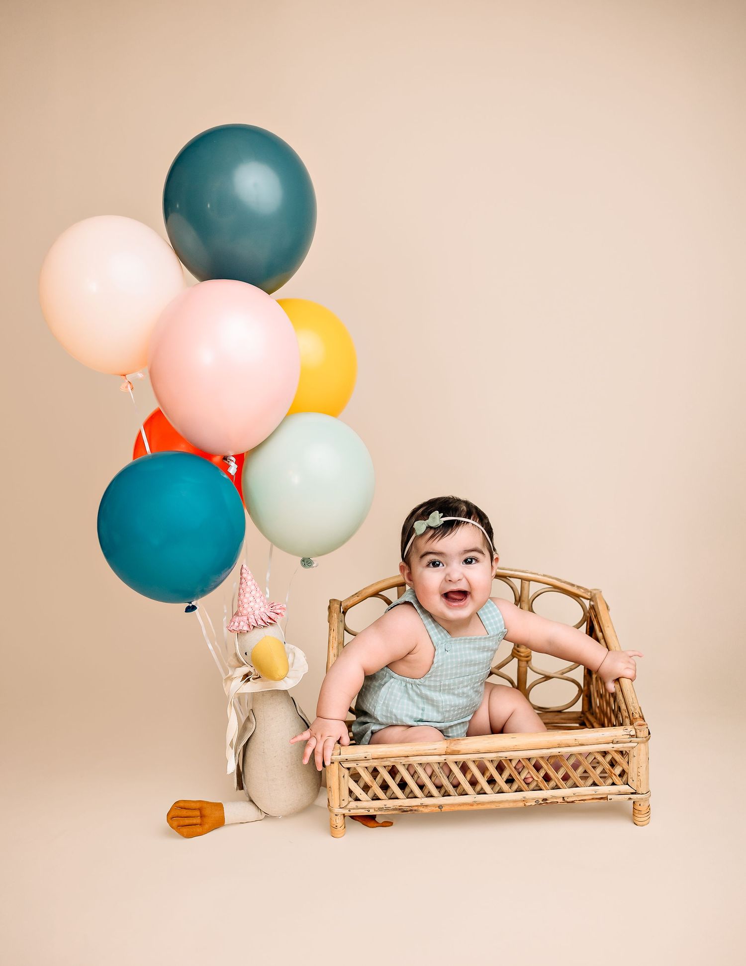 Cheerful baby photo session with colorful balloons and wicker furniture on beige backdrop.