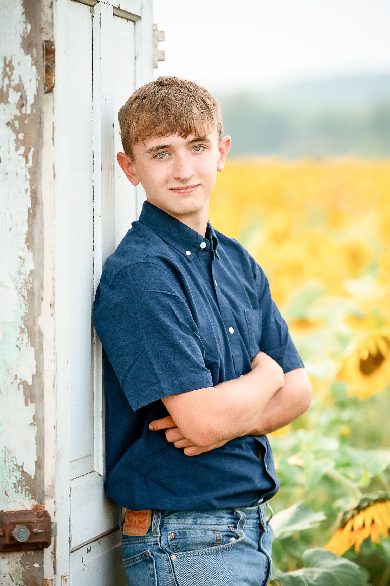 A young person in a navy polo shirt poses against a white door with a sunflower field in the background.