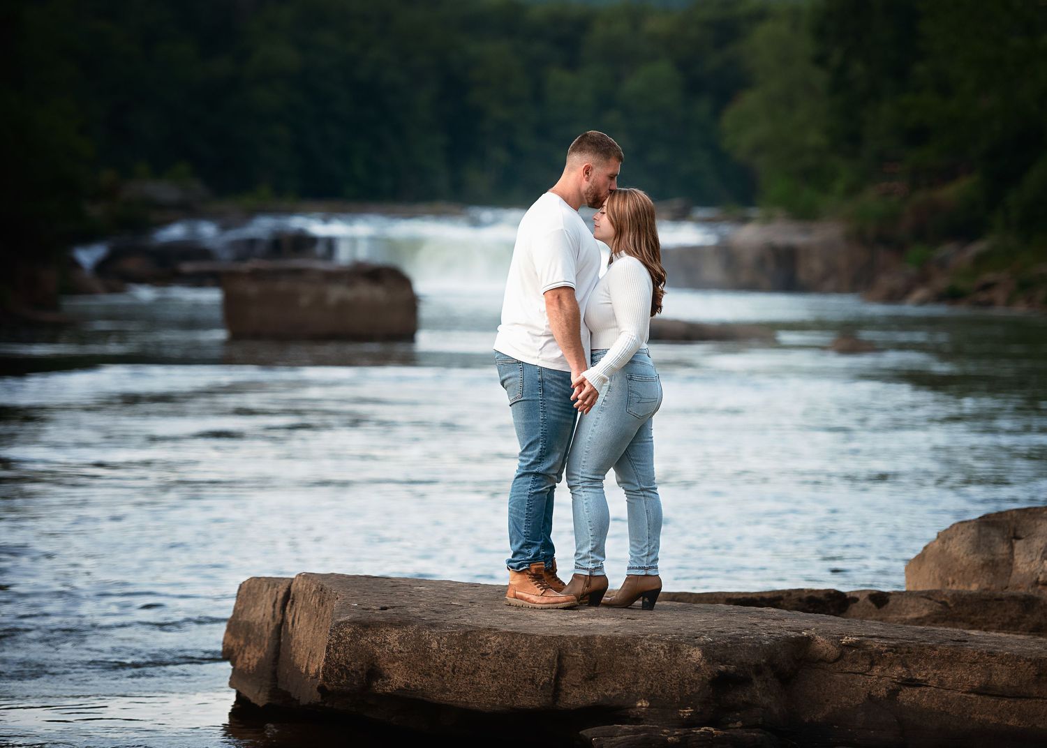 A couple embraces while standing on rocks at the edge of a rushing river with cascading waterfalls in the background.