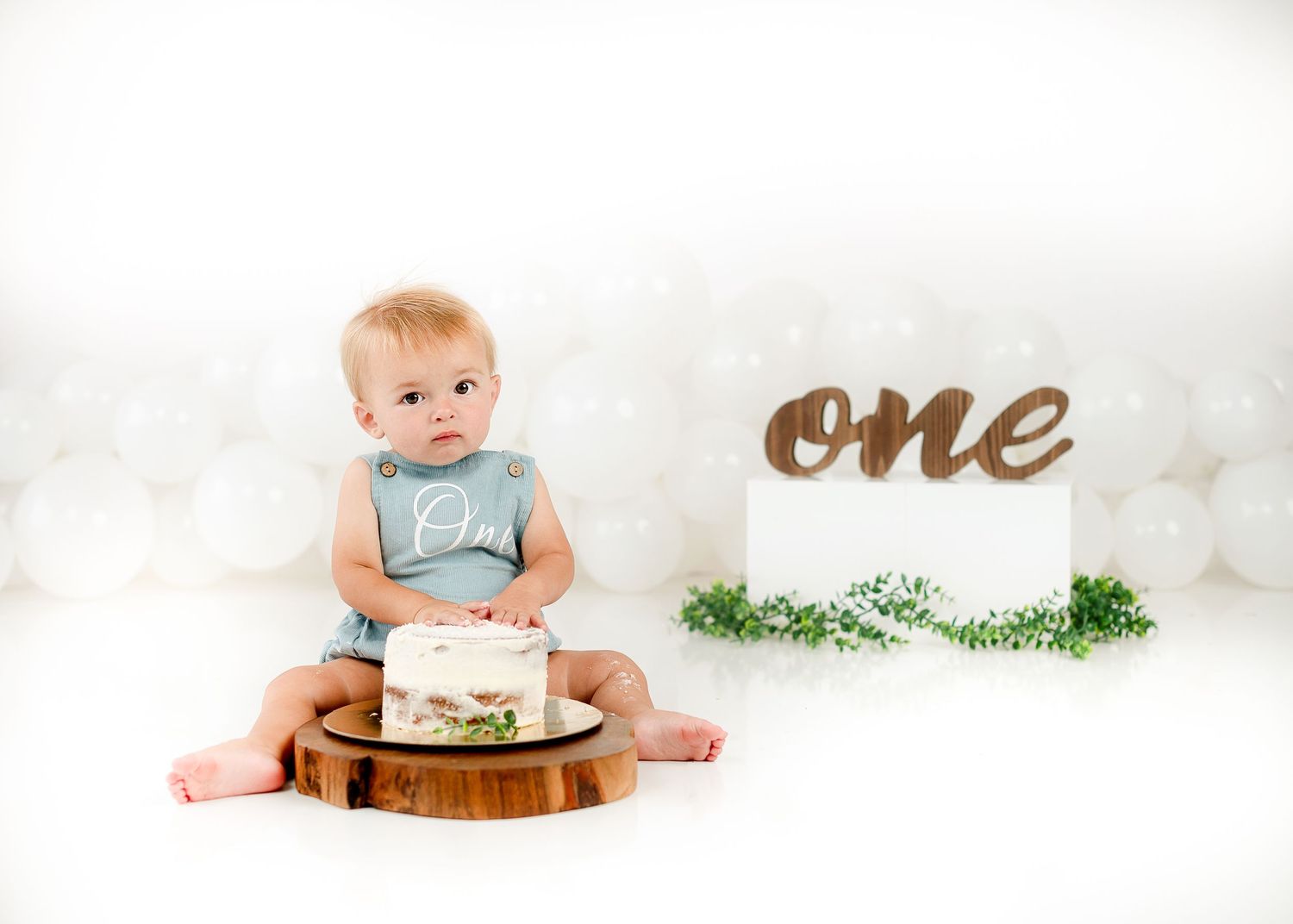 Minimalist first birthday setup with wooden cake stand and greenery on white background.
