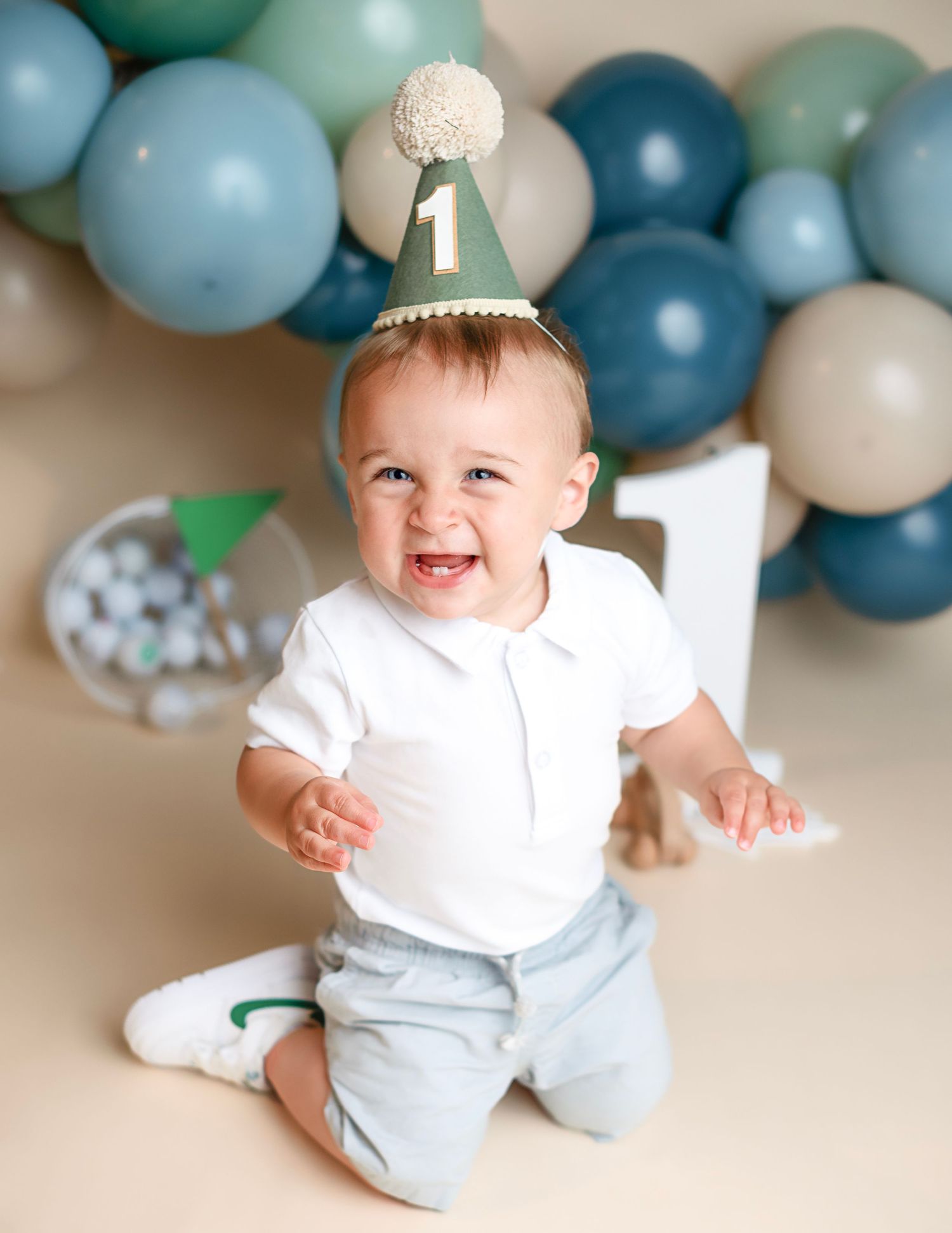 Baby in party hat kneeling with blue and white balloon backdrop for first birthday photo.