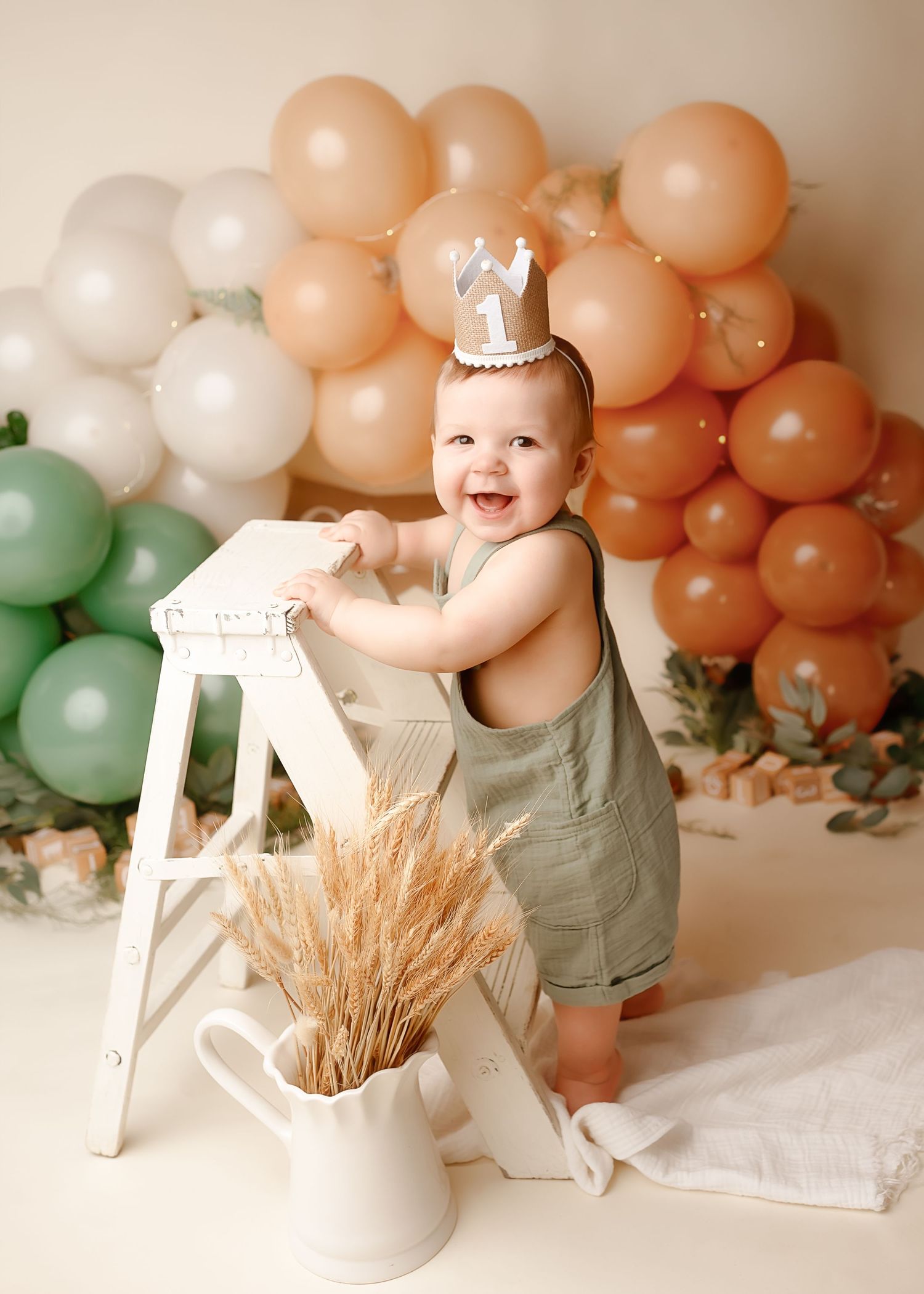 Baby in sage romper standing by white ladder with balloon arch backdrop.