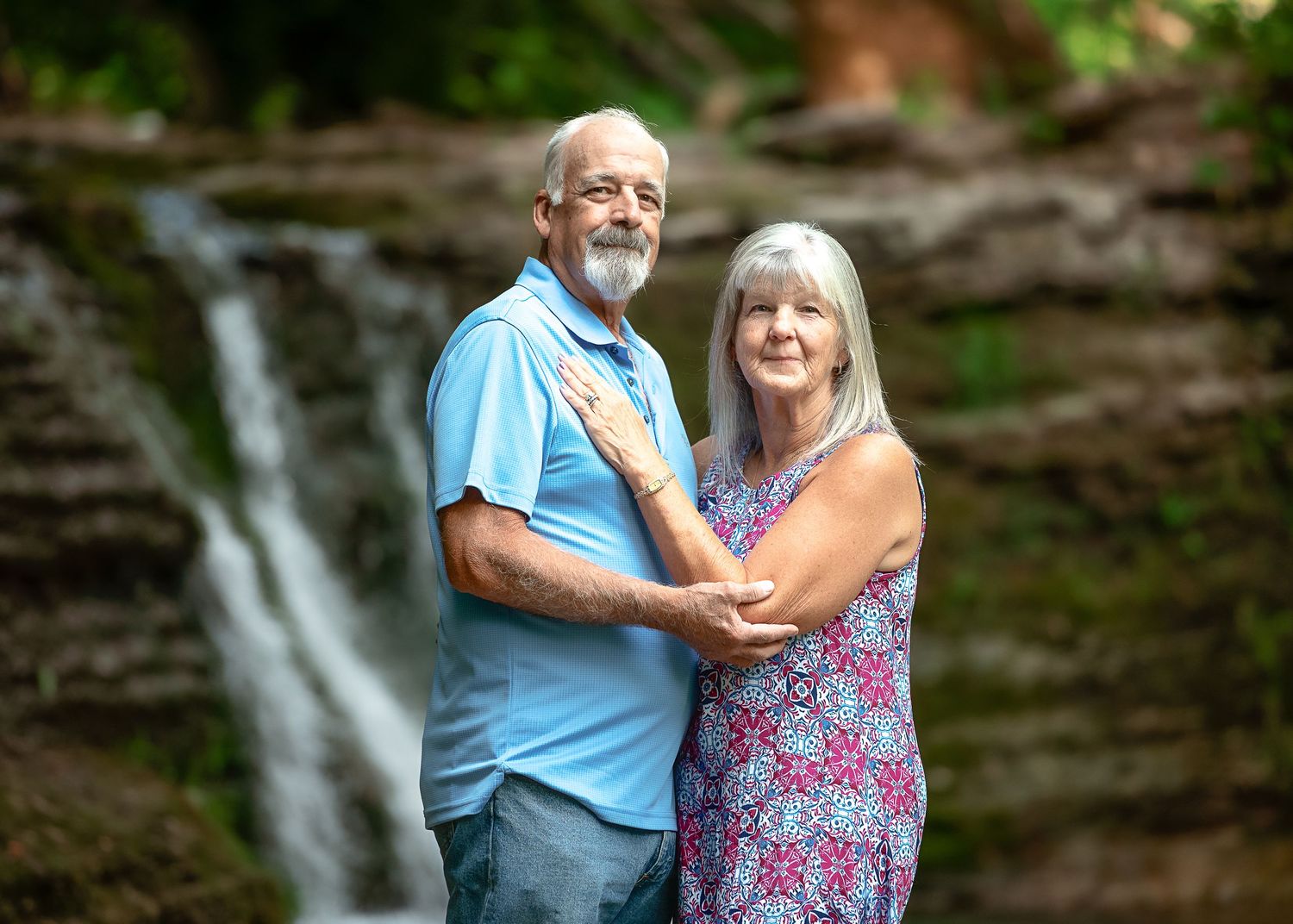 A couple embraces near a scenic waterfall in a lush forest setting.